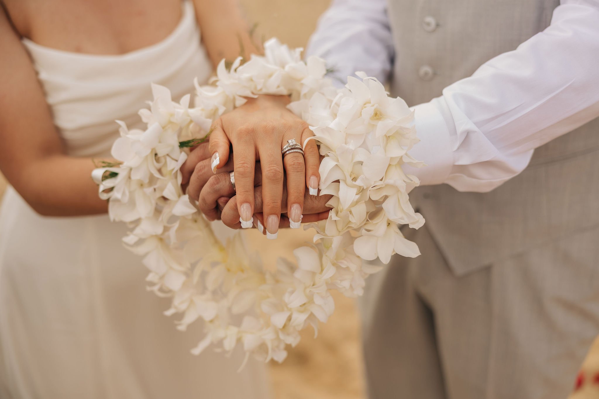 Close-up of a bride and groom holding hands, with the bride wearing rings and a floral wristband, during a wedding ceremony.