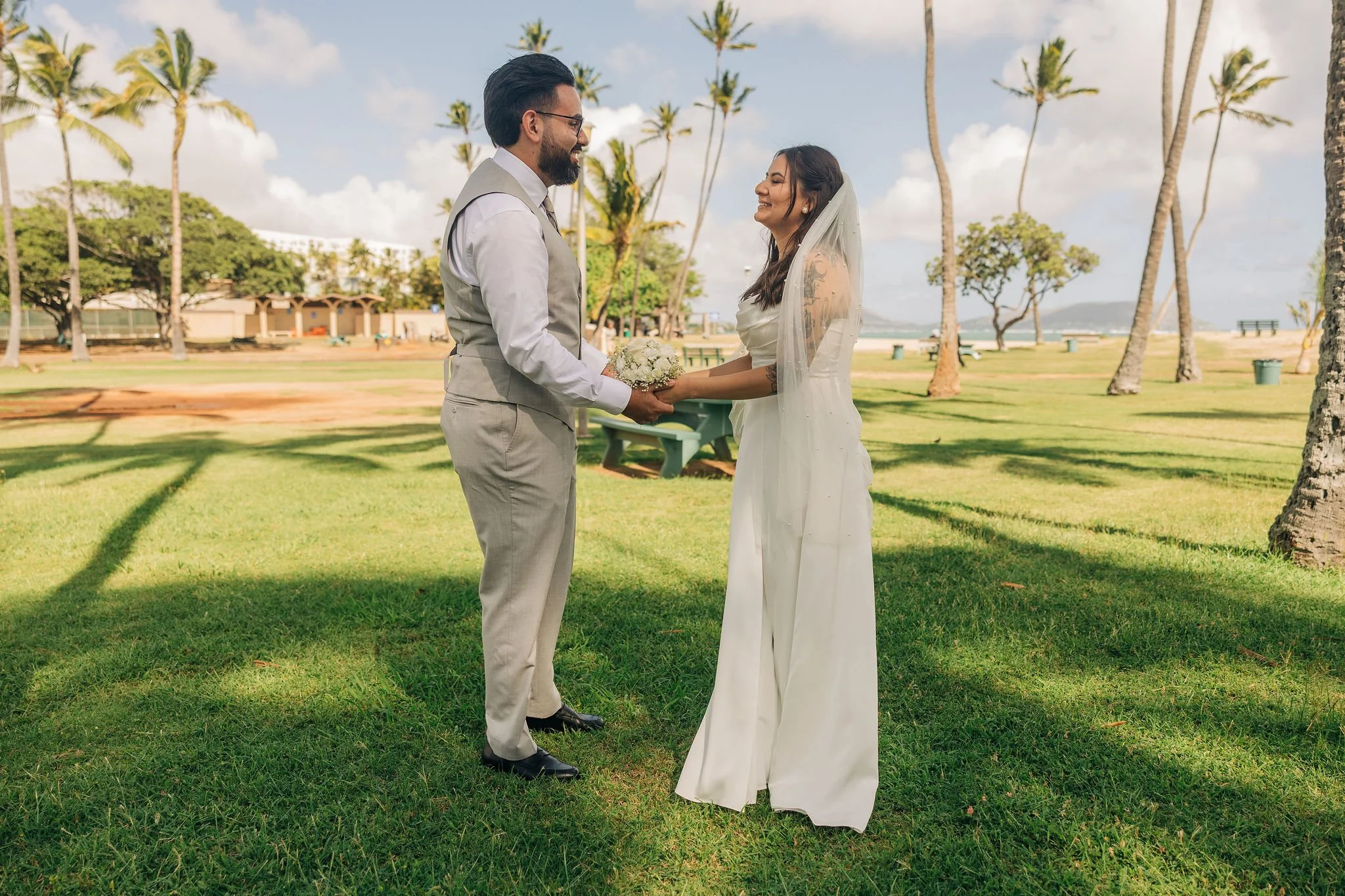 A bride and groom standing on grass holding hands and exchanging a bouquet at an outdoor wedding with palm trees and a beach background.