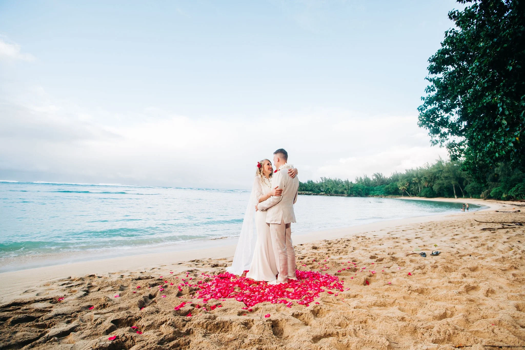 A bride and groom in wedding attire embracing on a sandy beach, surrounded by pink flower petals with the ocean and lush green trees in the background.