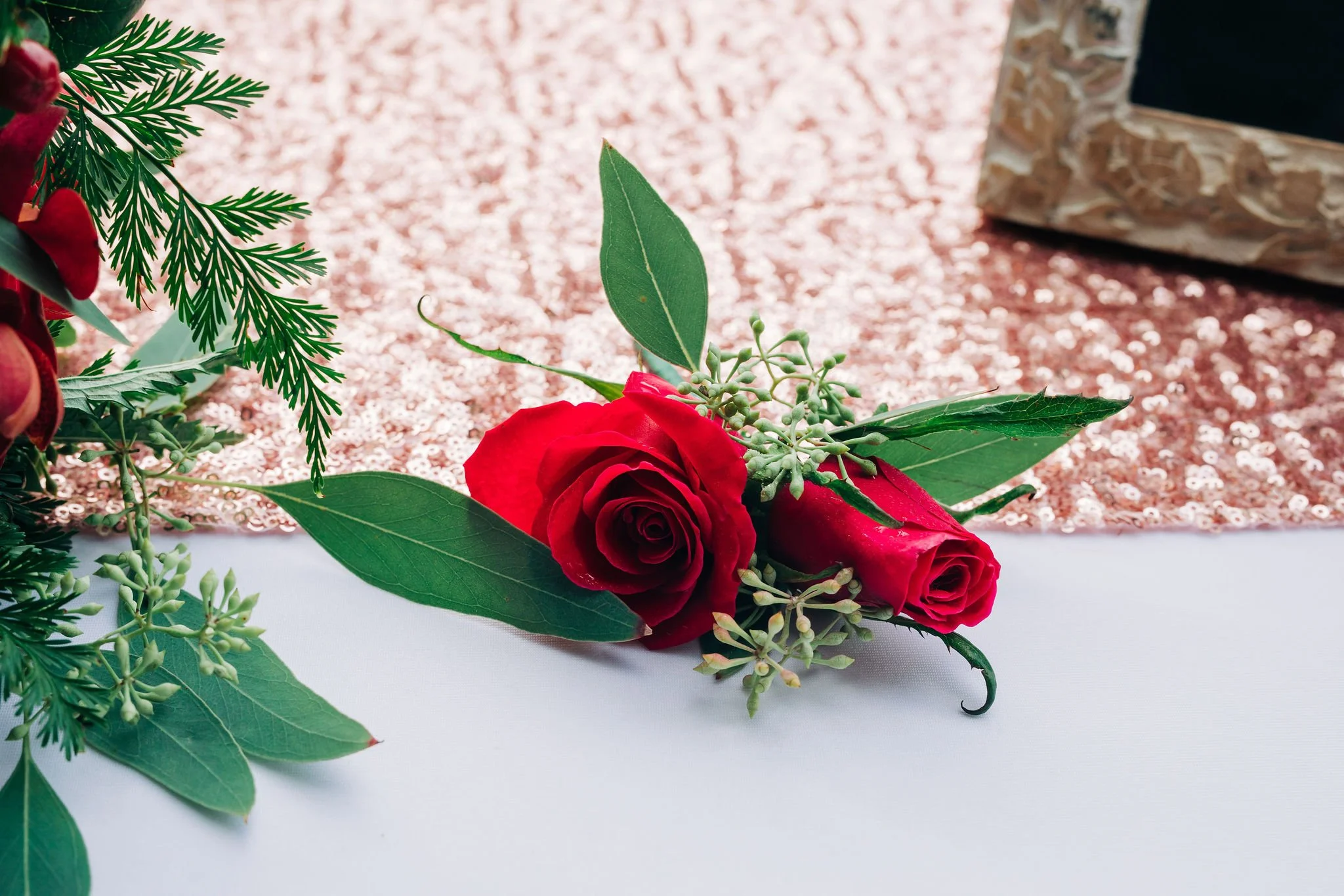 Red roses with green leaves and small white flowers on a white table with a pink sequin and beige patterned background.