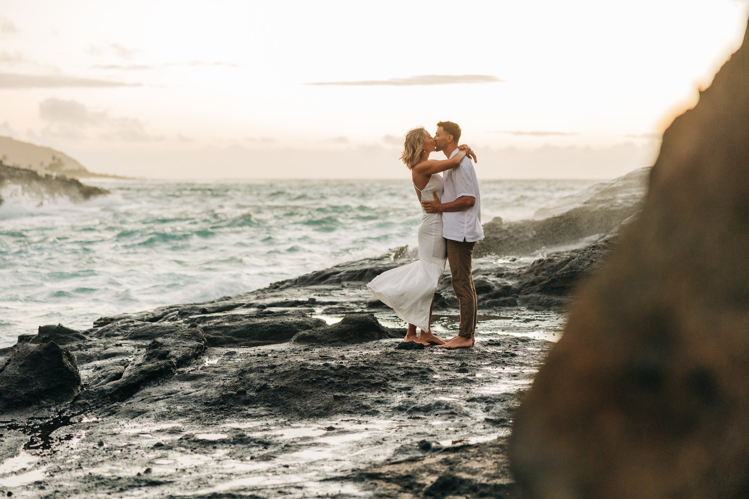 A couple in wedding attire embraces on rocky shoreline at sunset with ocean waves in the background.