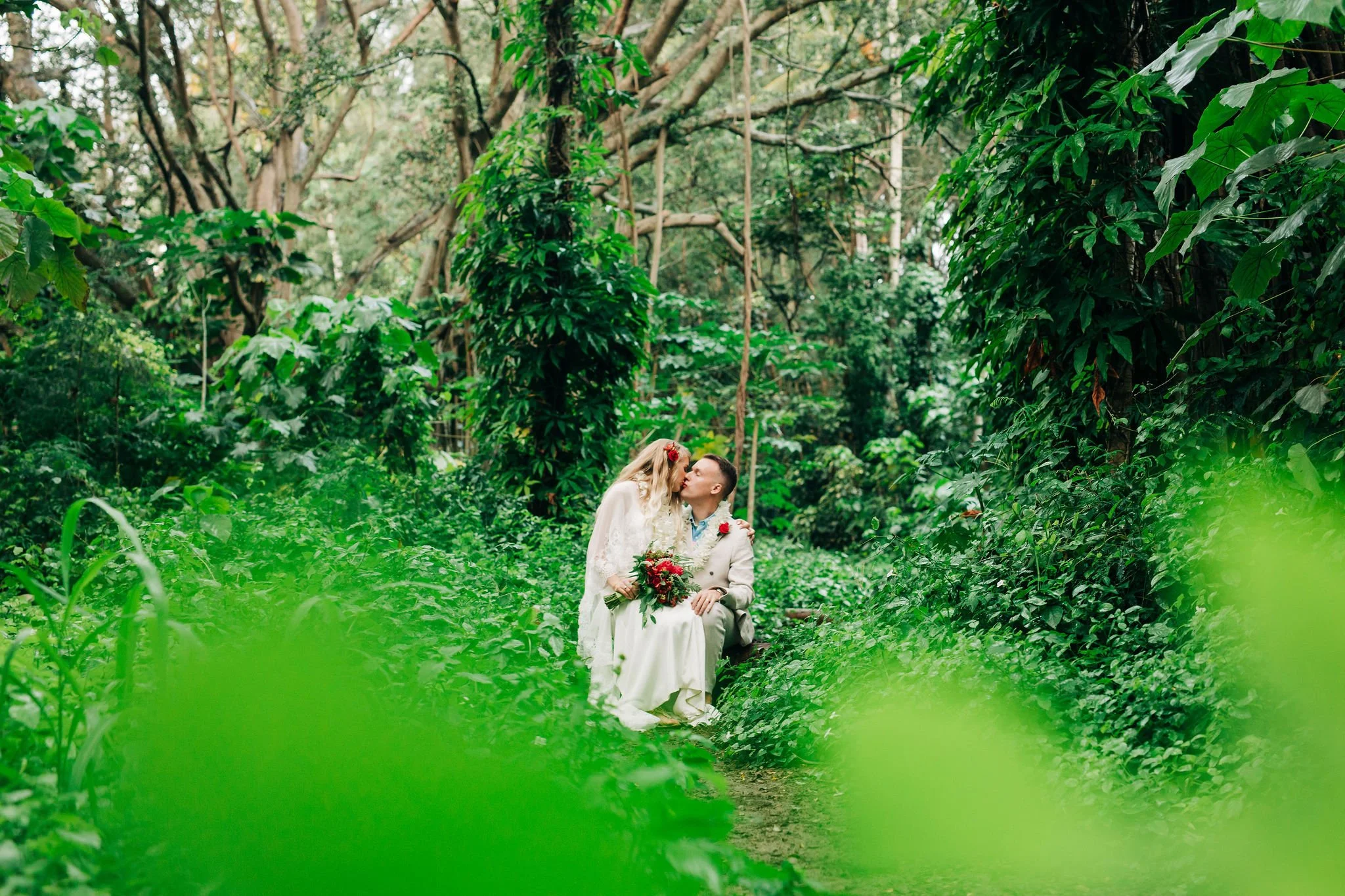 A bride and groom sharing a kiss in a lush green forest, surrounded by trees and dense foliage during a wedding photoshoot.