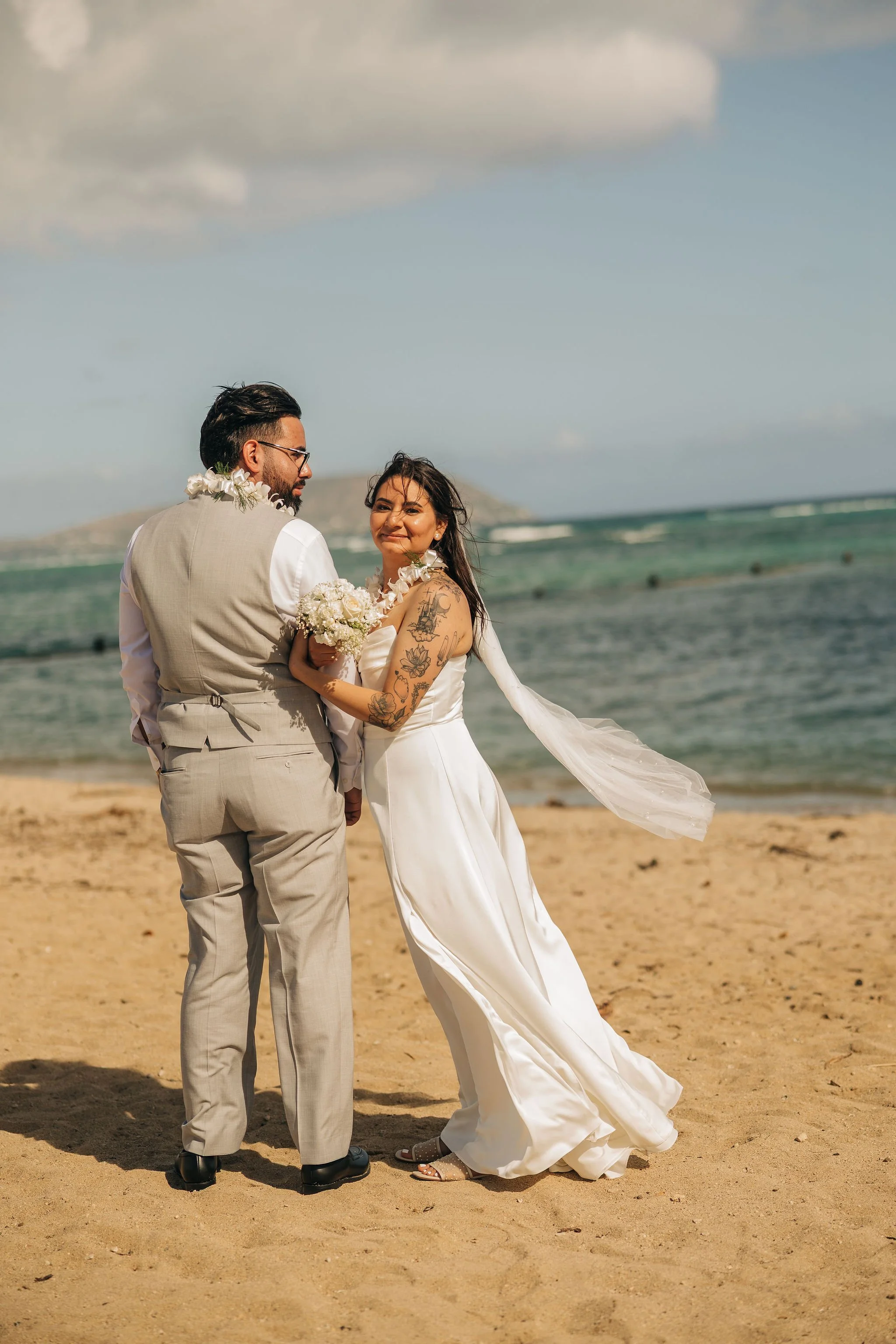 A couple in wedding attire standing on a sandy beach, with the ocean and a small island in the background. The bride holds a bouquet and smiles at the camera, while the groom looks at her.