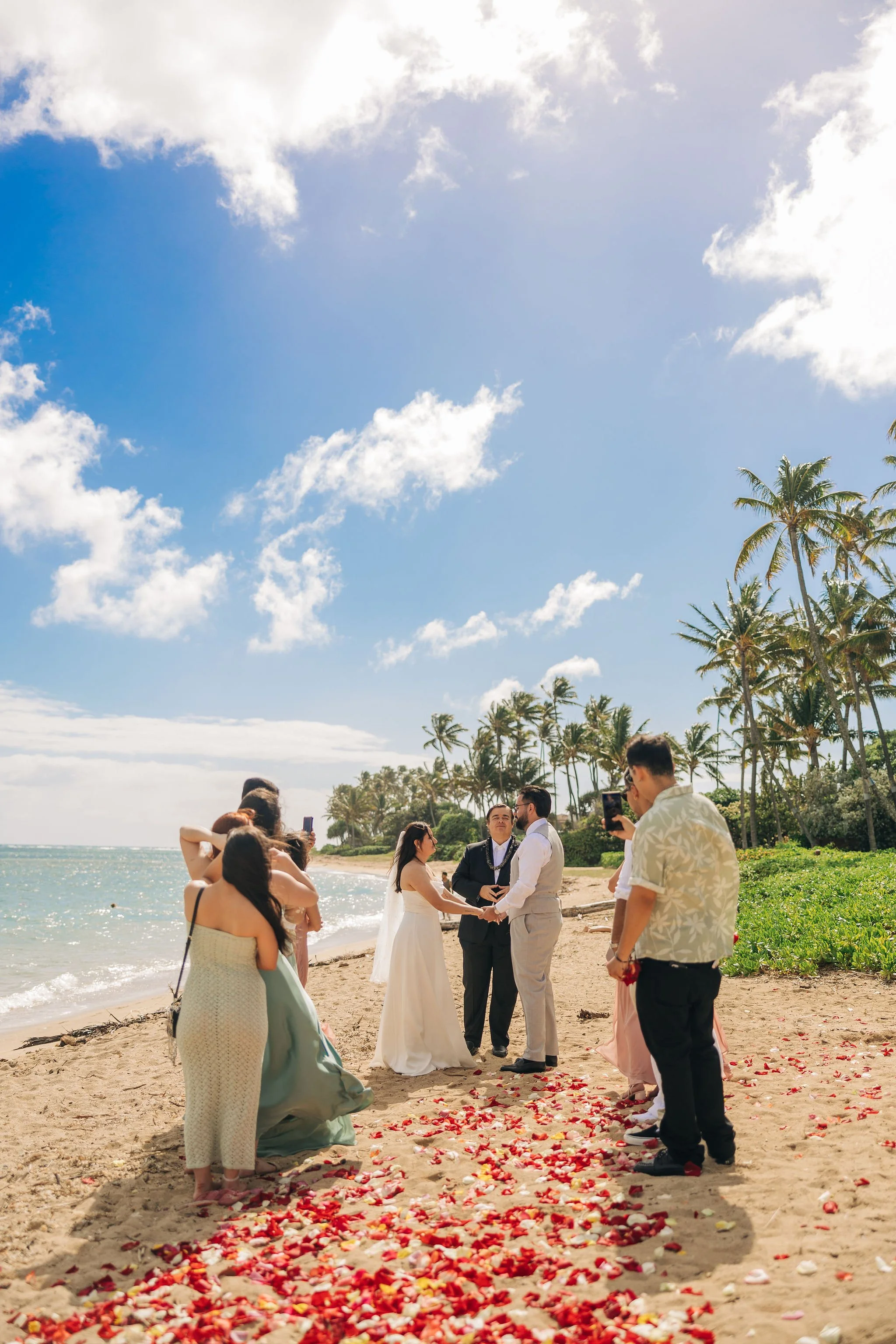 A beach wedding ceremony with a couple holding hands, officiant, bridesmaids, groomsmen, and guests on the sandy shore lined with palm trees on a bright, sunny day.
