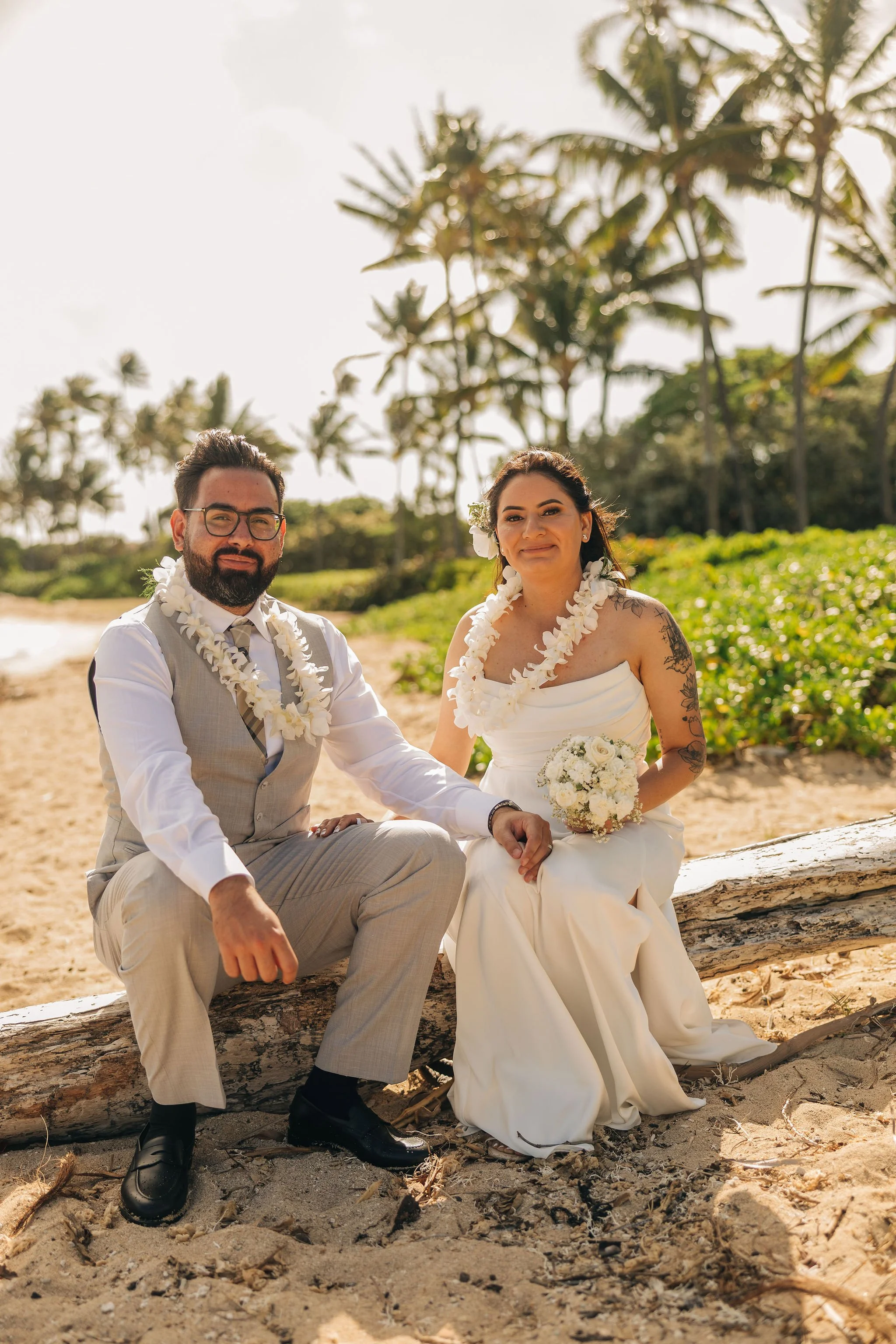 A couple sitting on a log on the beach, dressed in wedding attire with palm trees in the background, enjoying a sunny day.