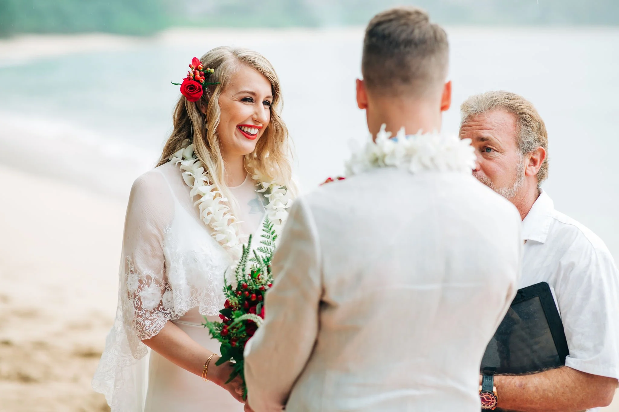 A smiling bride with a red flower in her hair, wearing a white lace dress and floral lei, holds a bouquet of red and green flowers at a beach wedding, with an officiant and groom in white shirts.