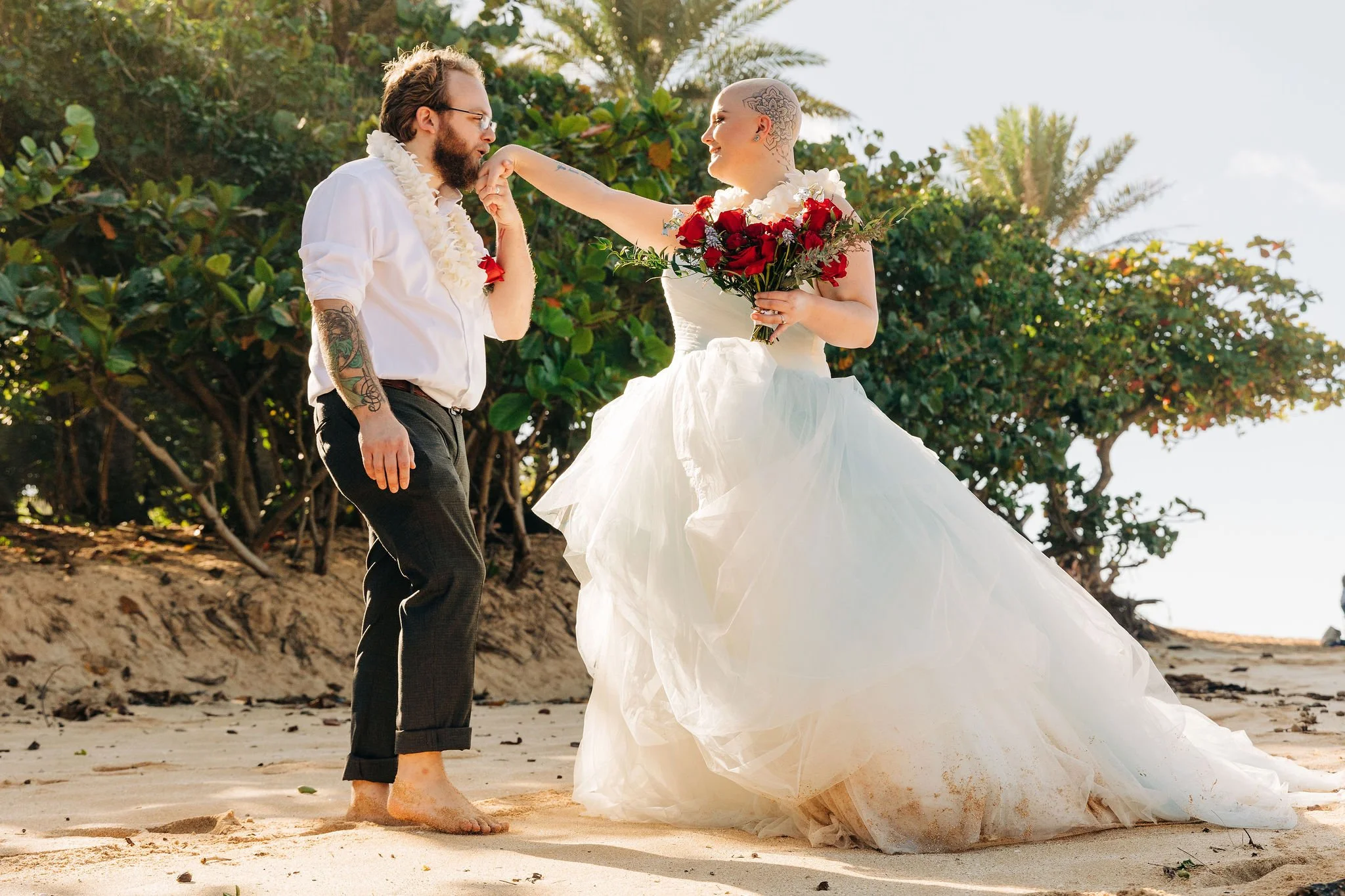 A couple on a beach, with the bride in a white wedding dress holding a bouquet of red roses, and the groom in black pants and a white shirt, with the groom kissing the bride's hand.