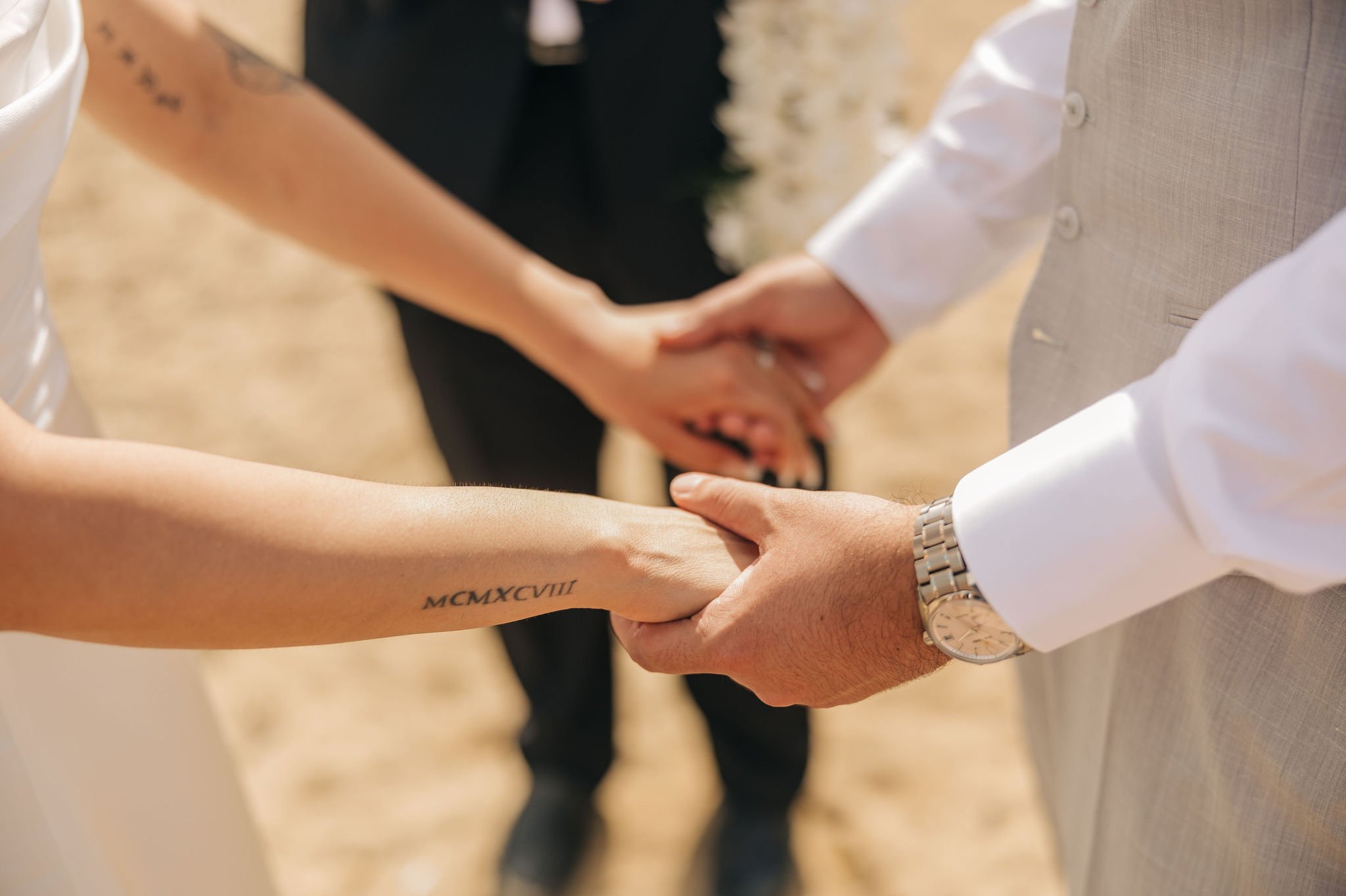 A couple holding hands during their wedding ceremony, with the bride's tattoo on her forearm and a wedding ring visible. The groom is wearing a light-colored suit and a watch.