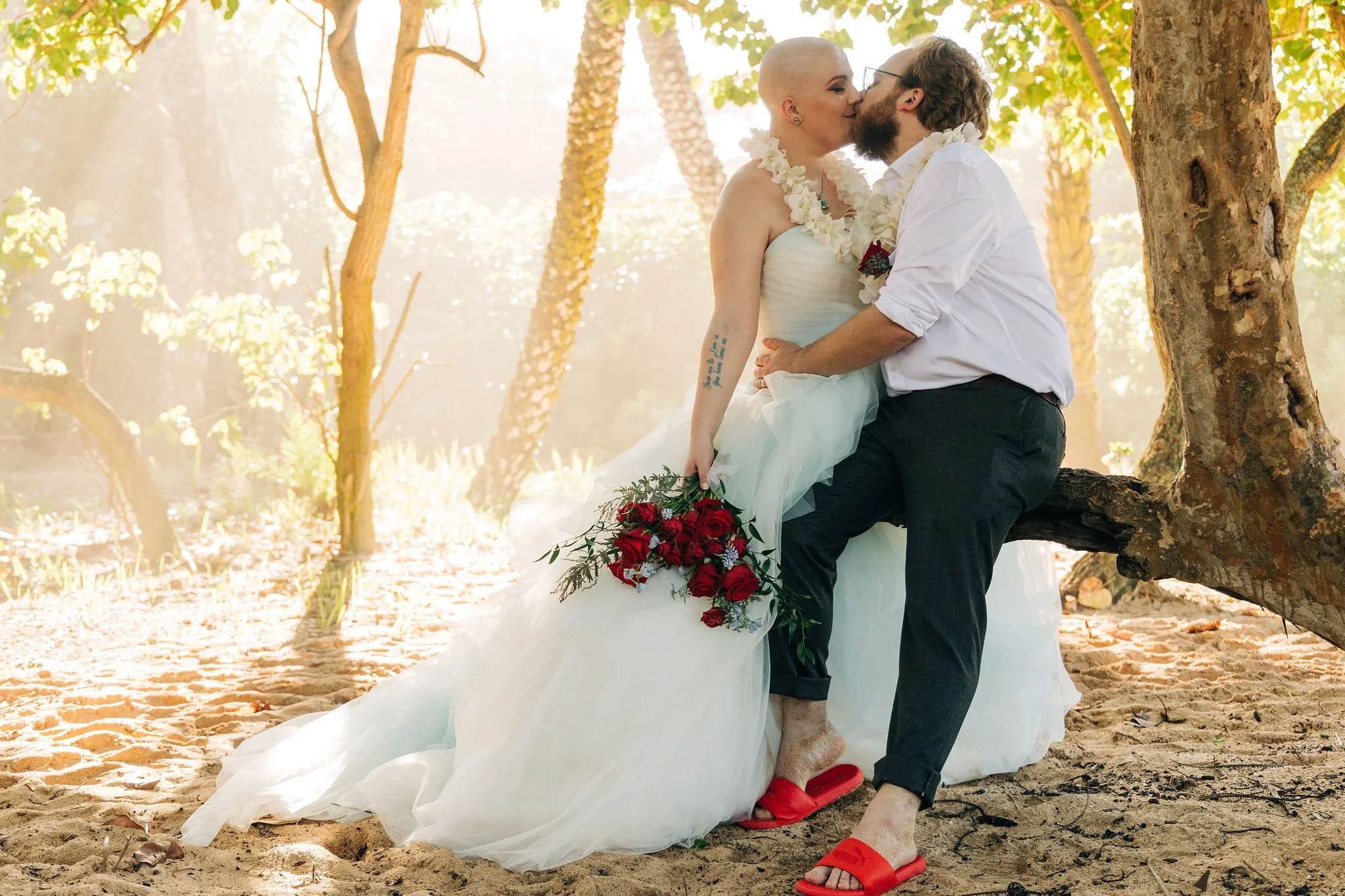 A couple is sitting on a tree branch in a sunlit forest, sharing a kiss. The woman wears a wedding dress and lei, holds a bouquet of red roses, and has a tattoo on her arm. The man wears a white shirt, dark pants, and red slippers.