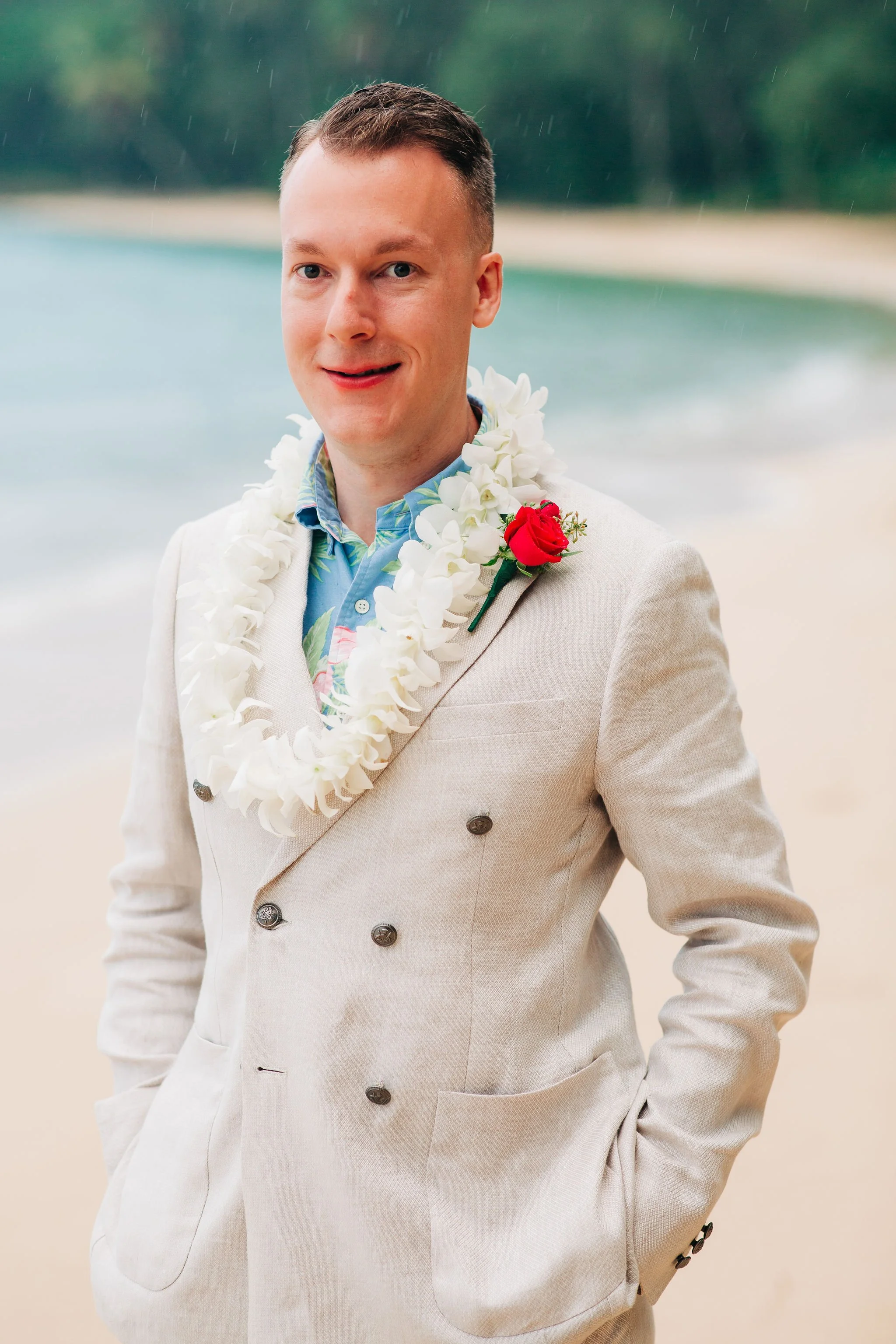 A man in a beige double-breasted suit jacket with a lei around his neck, standing on a beach in the rain. The background shows the ocean and a tree-lined shore.