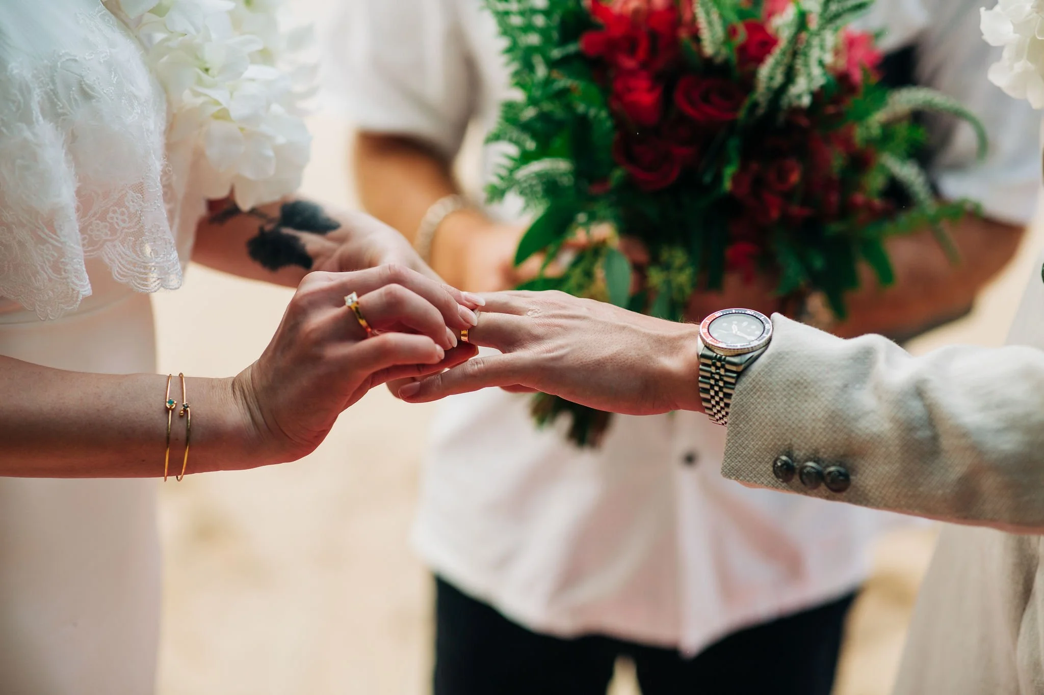 A couple exchanging wedding vows with a stand-in to the reception, with the bride placing a ring on the groom's finger, and a bouquet of red roses in the background.