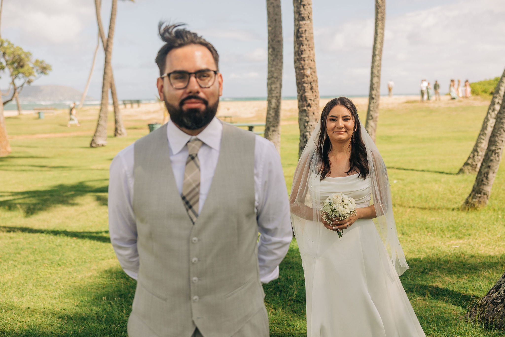 A man in glasses and a vest stands in the foreground of a scenic outdoor wedding photo, with a woman in a white wedding dress holding a bouquet of flowers in the background, set in a grassy park with palm trees, and the ocean visible in the distance.