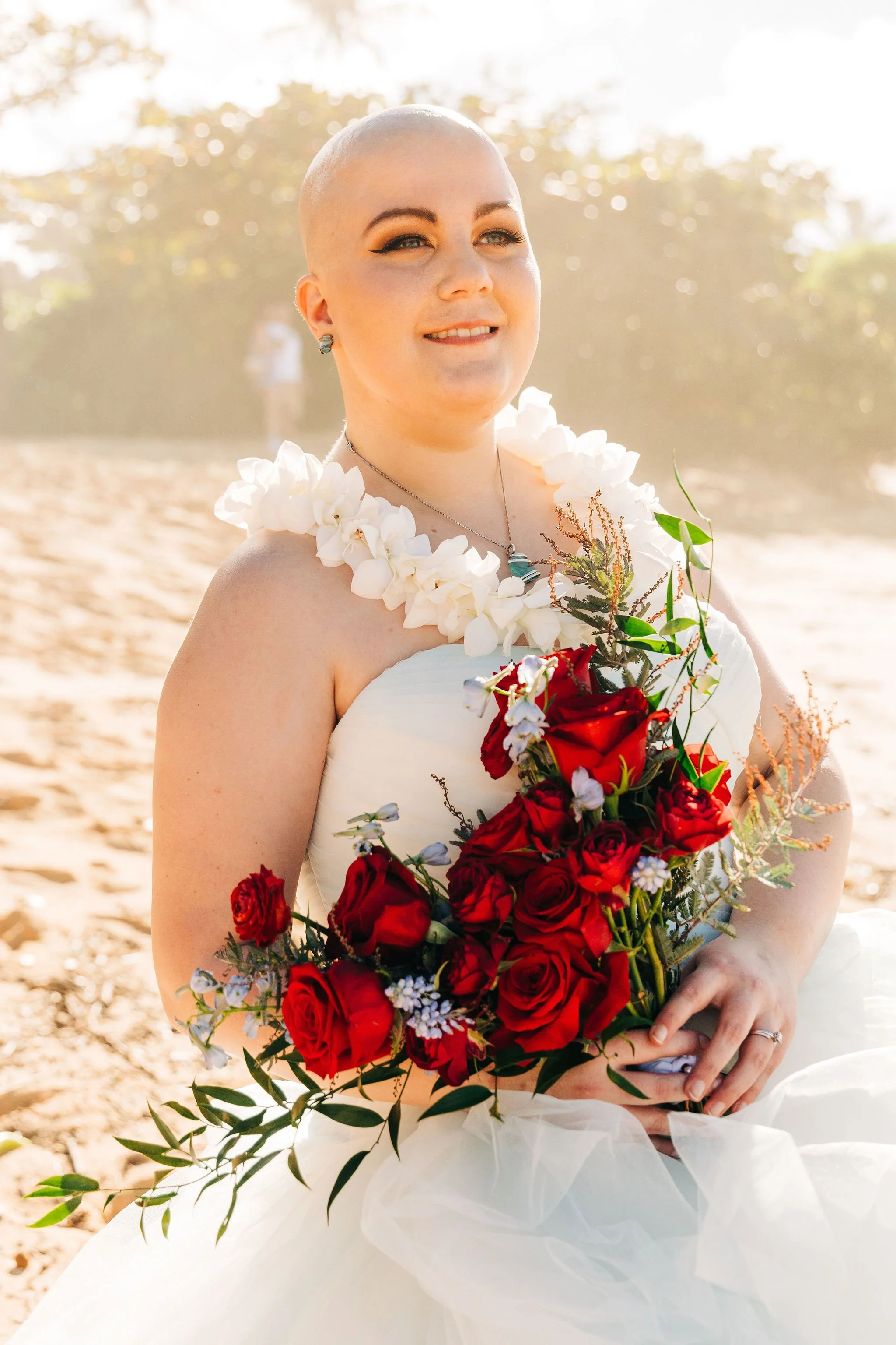 A woman with a shaved head is holding a large bouquet of red roses and greenery, wearing a strapless white dress, standing outdoors on a sunny day with trees in the background.