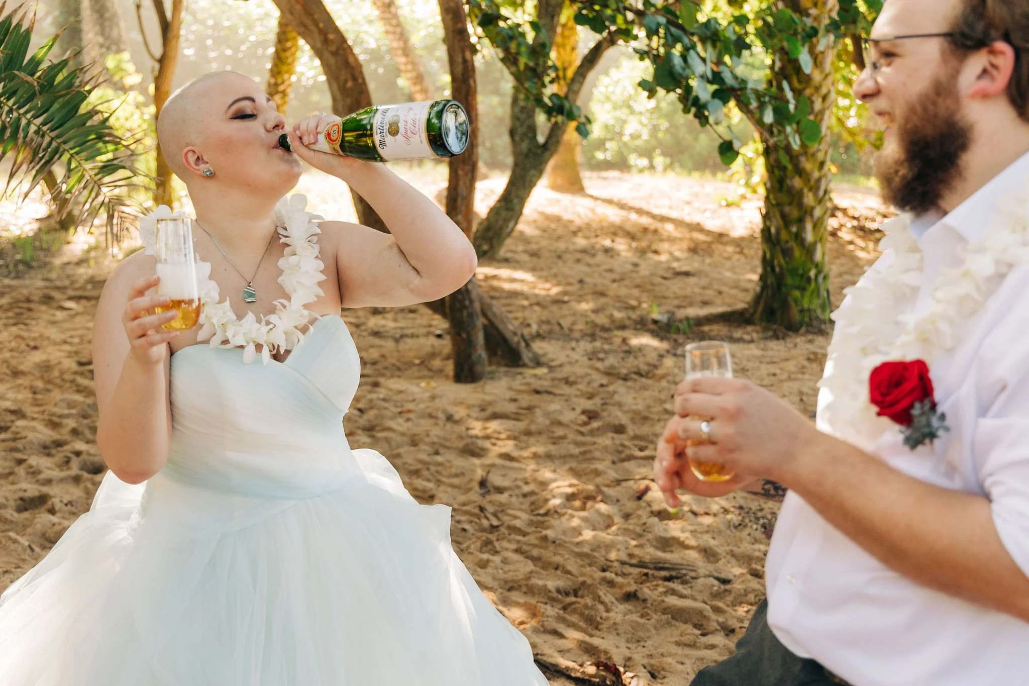 A bride with a shaved head and a white dress takes a shot of beer while holding a glass of beer, and a groom with glasses and a beard, dressed in a white shirt with a red rose boutonniere, stands in front of her with a beer in hand in a wooded outdoo