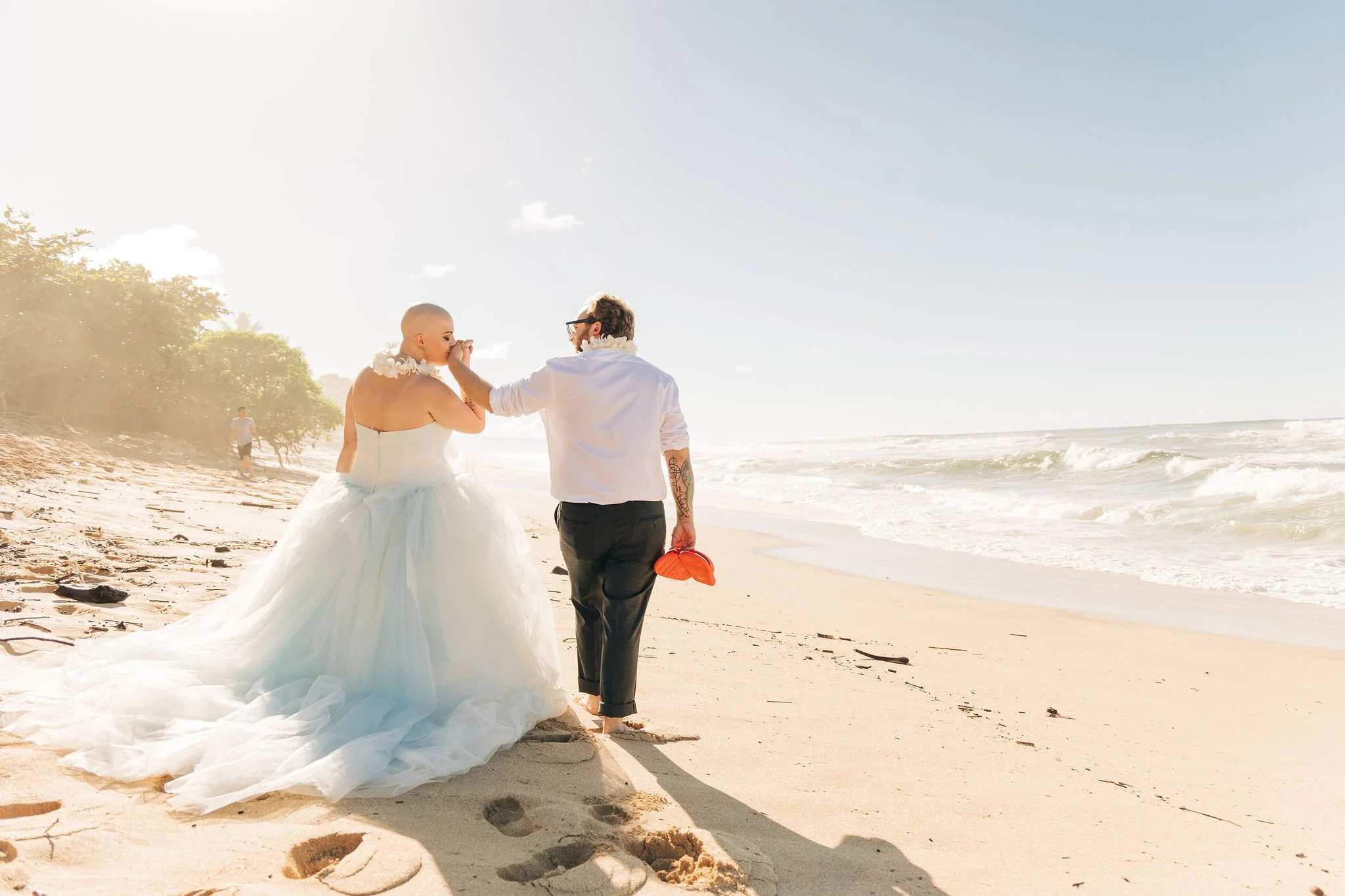 A person in a wedding dress and a person in casual clothing walking along a sandy beach with ocean waves, trees, and a clear sky in the background.