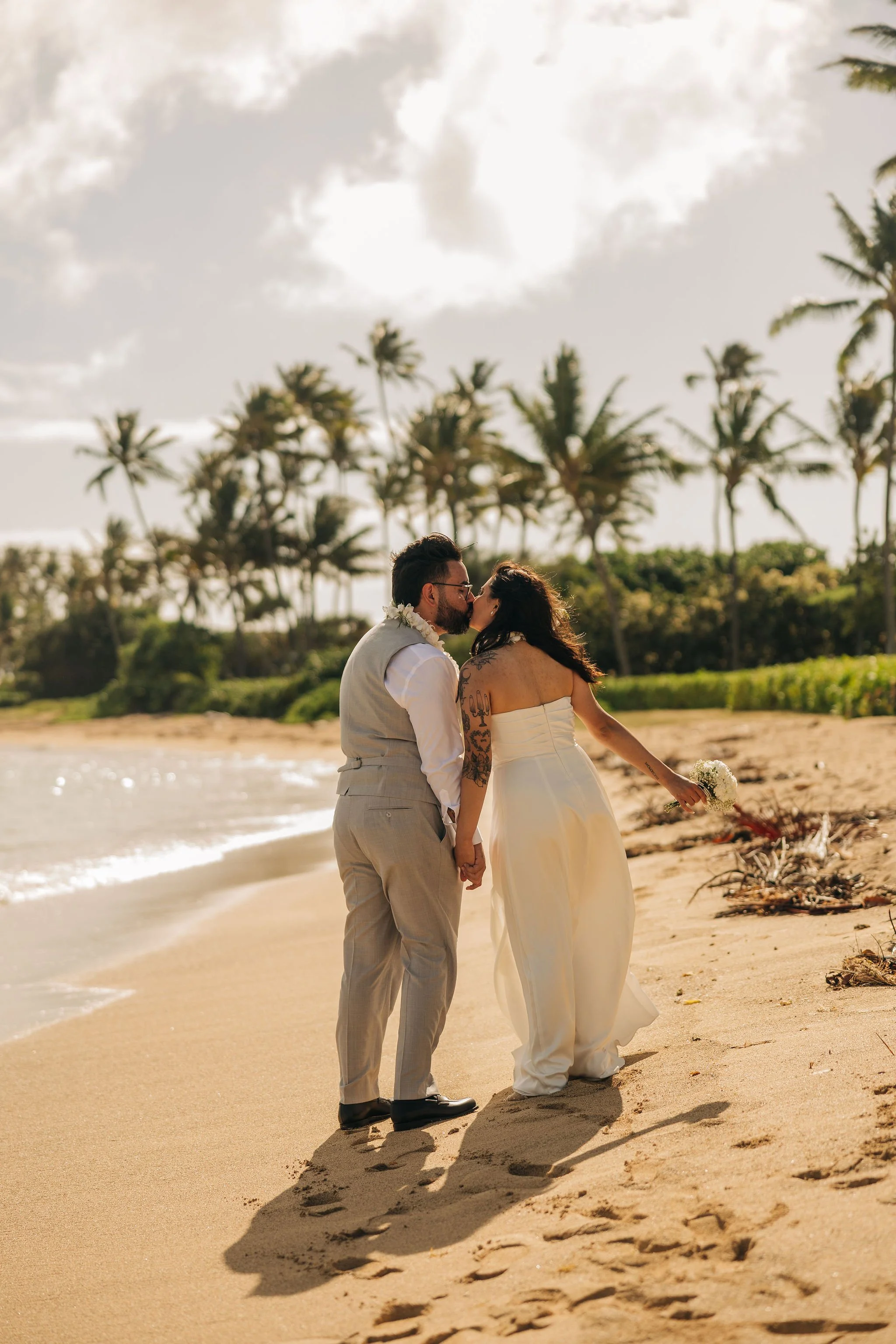 A newlywed couple sharing a kiss and holding hands on a beach with palm trees in the background, during sunset.