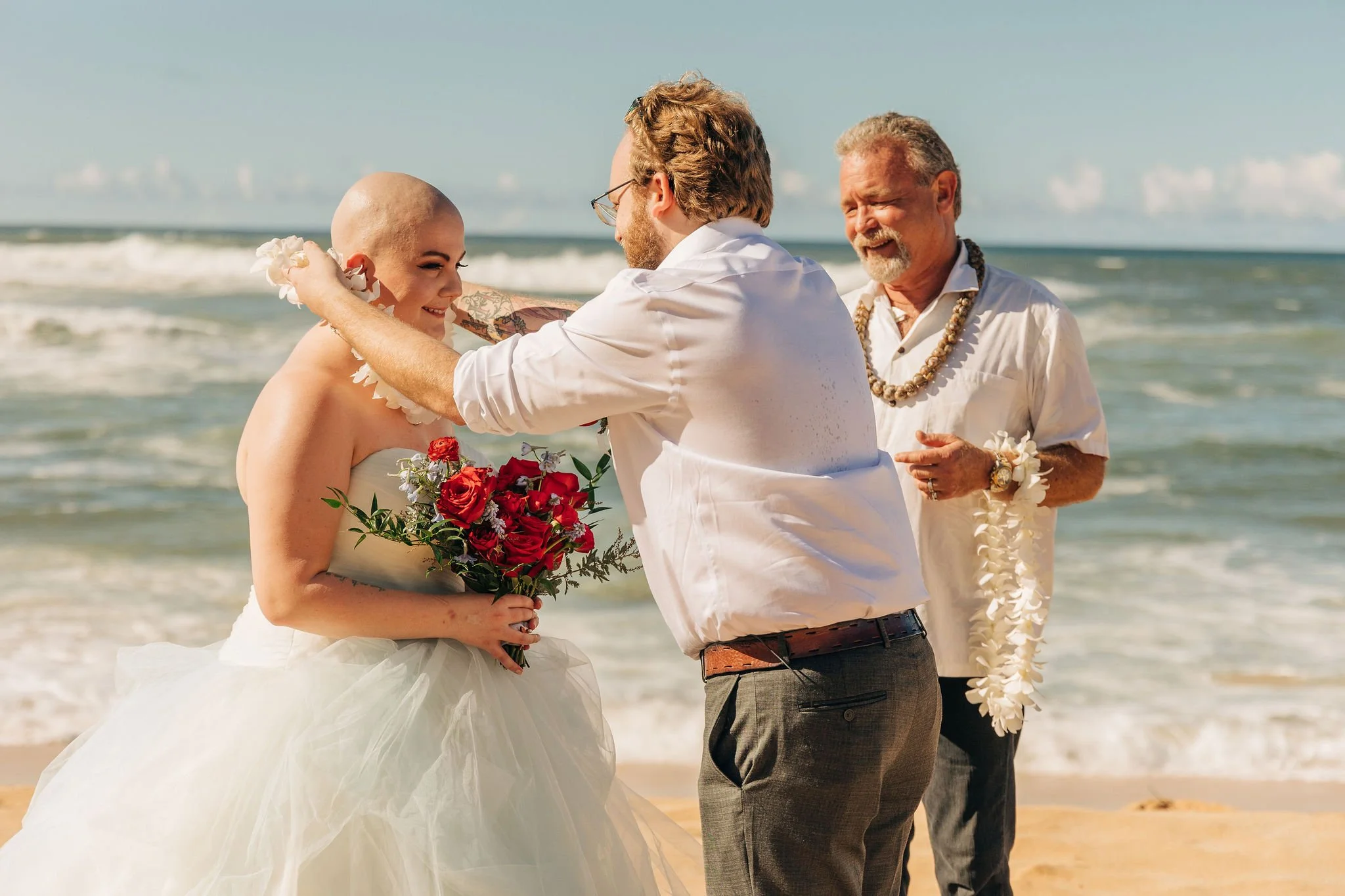 A wedding ceremony taking place on a beach, with the bride holding a bouquet of red roses, the groom adjusting her hair, and an officiant smiling behind them.