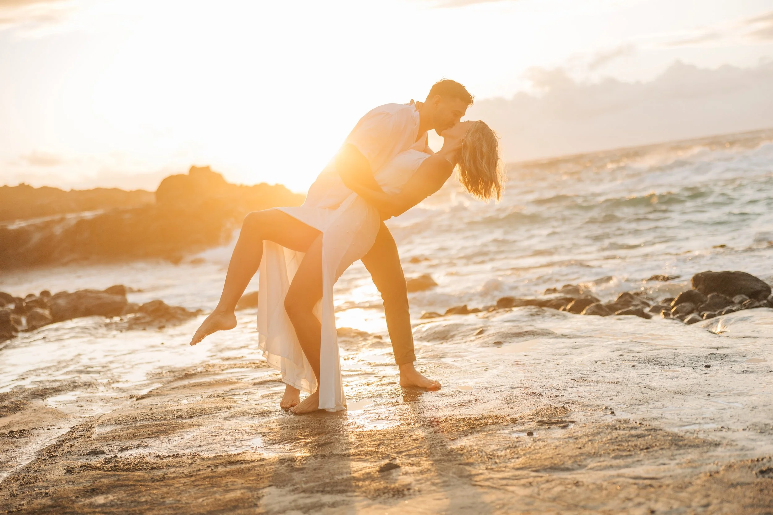 A couple dancing on the beach at sunset, with the man dipping the woman while holding her around the waist, and the woman holding his shoulders, wearing light-colored clothing.