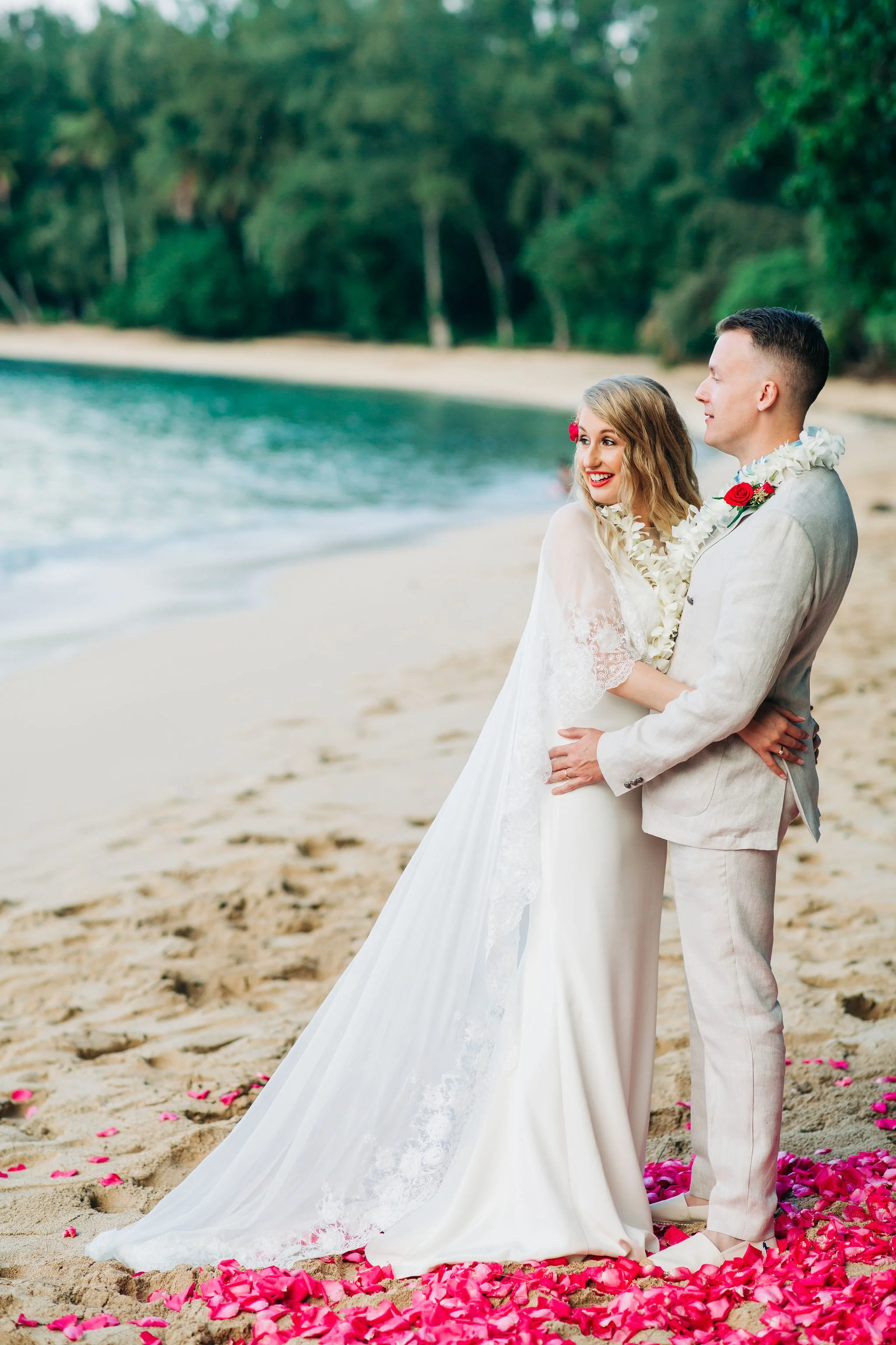 A bride and groom standing on a sandy beach, smiling and embracing, with pink flower petals on the ground and ocean in the background.