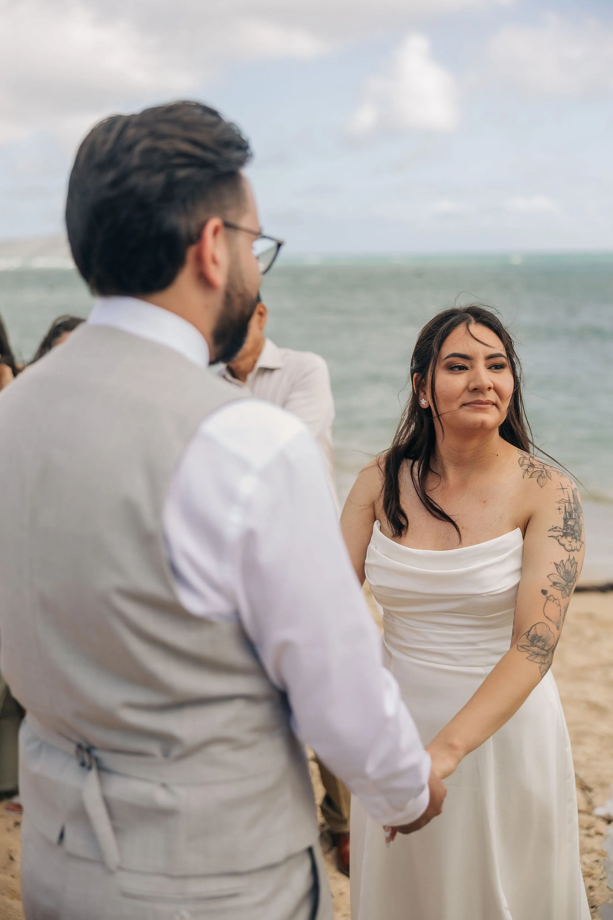 A bride and groom holding hands during a beach wedding ceremony, with the ocean and cloudy sky in the background.