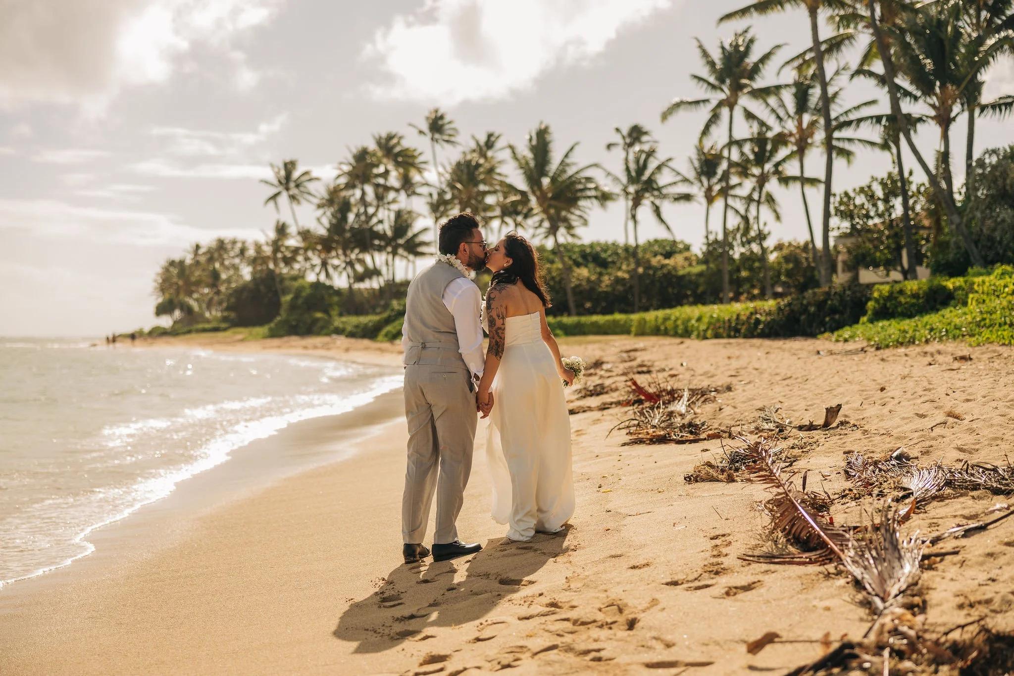 A couple dressed in wedding attire sharing a kiss on a beach with palm trees in the background during sunset.