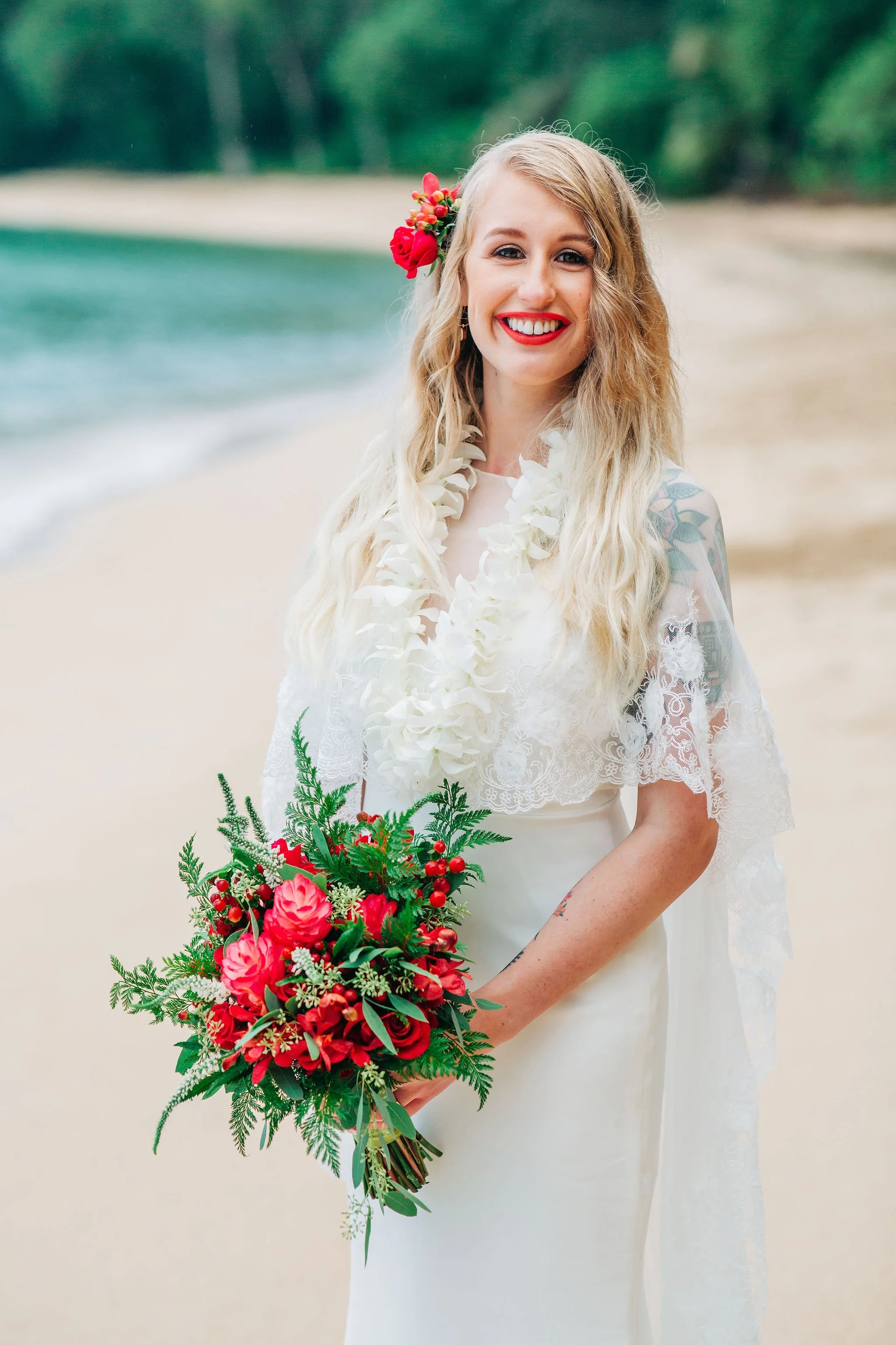 A smiling woman dressed in a wedding dress holding a bouquet of red flowers on a beach with sand and the ocean in the background.