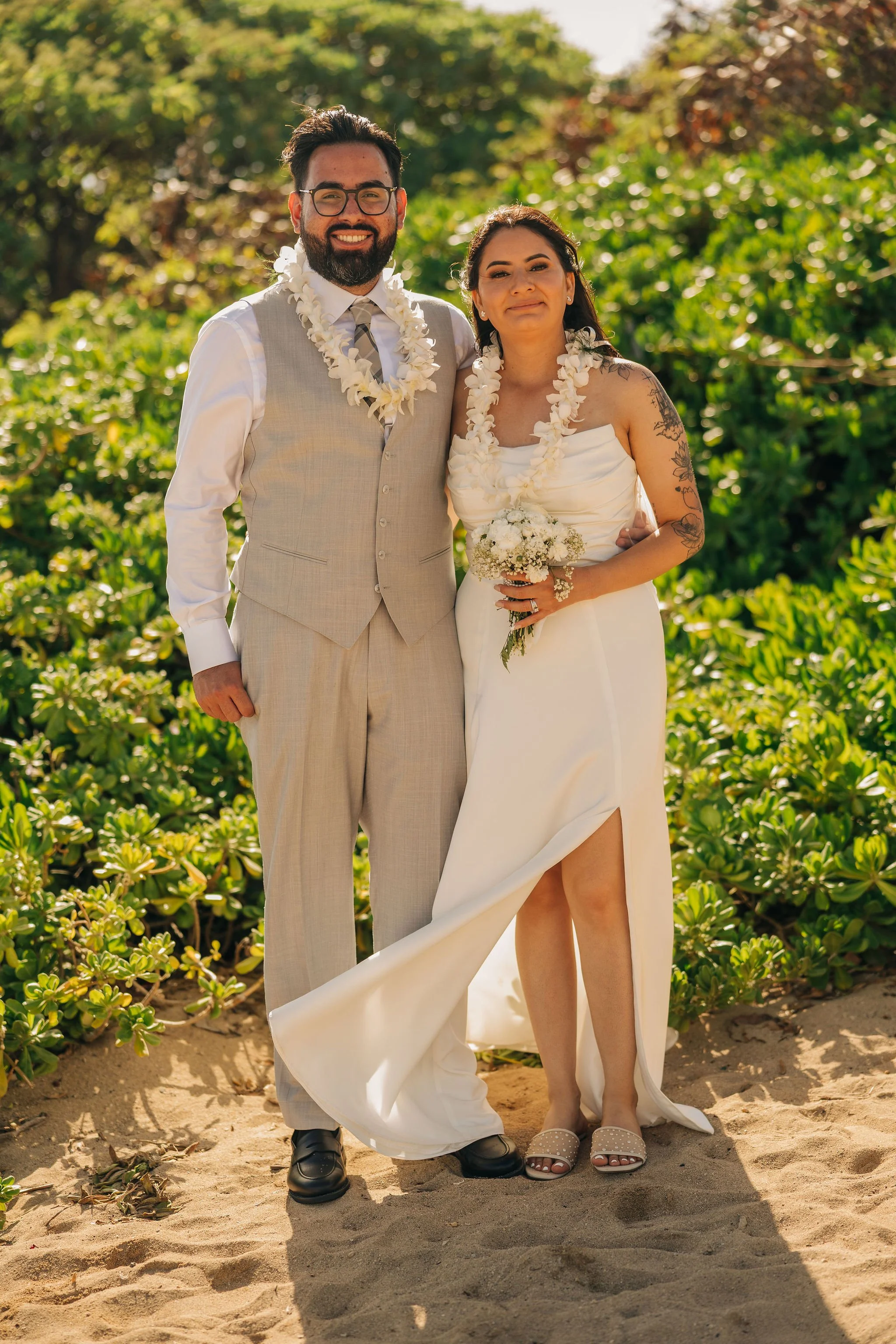 A couple in wedding attire standing on a sandy beach, surrounded by green bushes and trees, smiling at the camera. The man is wearing a beige suit vest with dress shirt and tie, glasses, and black shoes. The woman is wearing a white strapless wedding