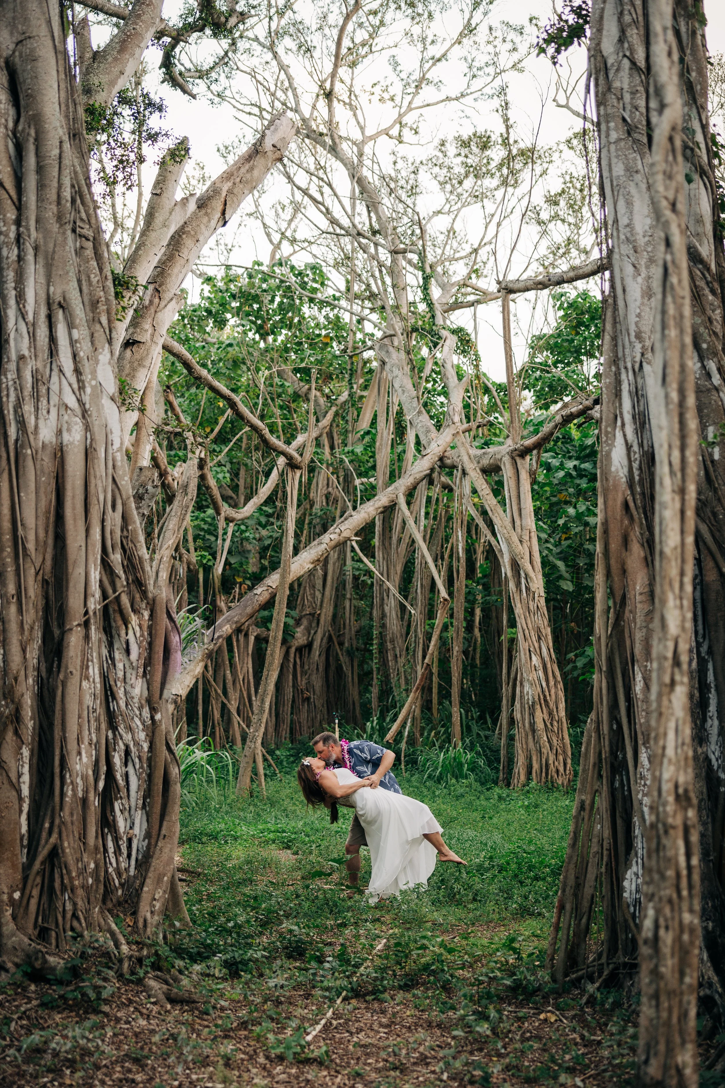 A couple in wedding attire dancing in a forest with large, twisting tree roots and greenery.