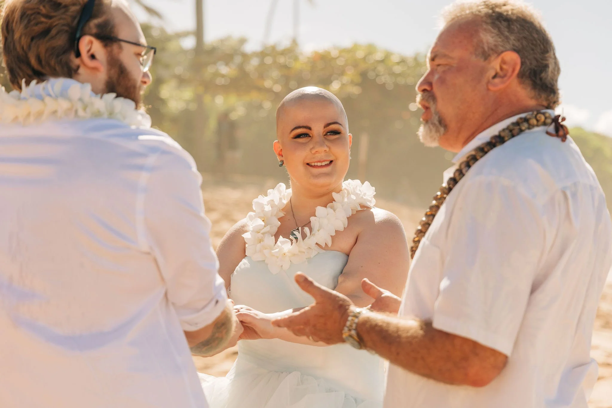 A couple getting married outdoors with a woman who has a shaved head, and a man with a beard and glasses. They are all wearing white and flower leis, standing on a sunny beach with trees in the background.