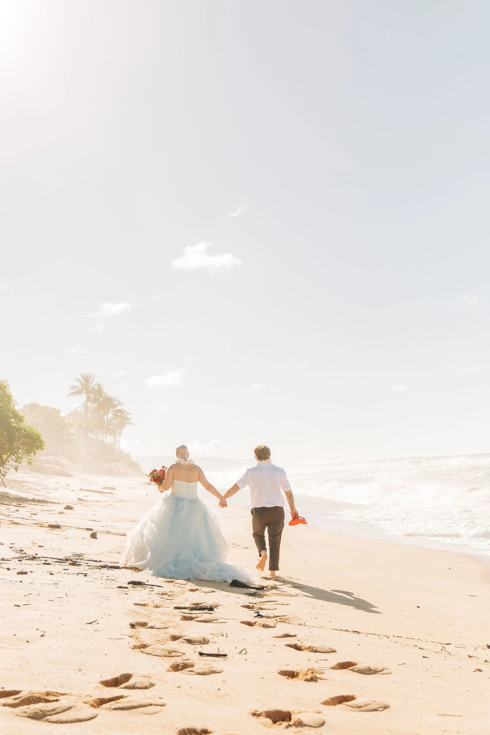 A bride and groom walking hand in hand along the beach at sunset, with footprints in the sand behind them and palm trees in the distance.