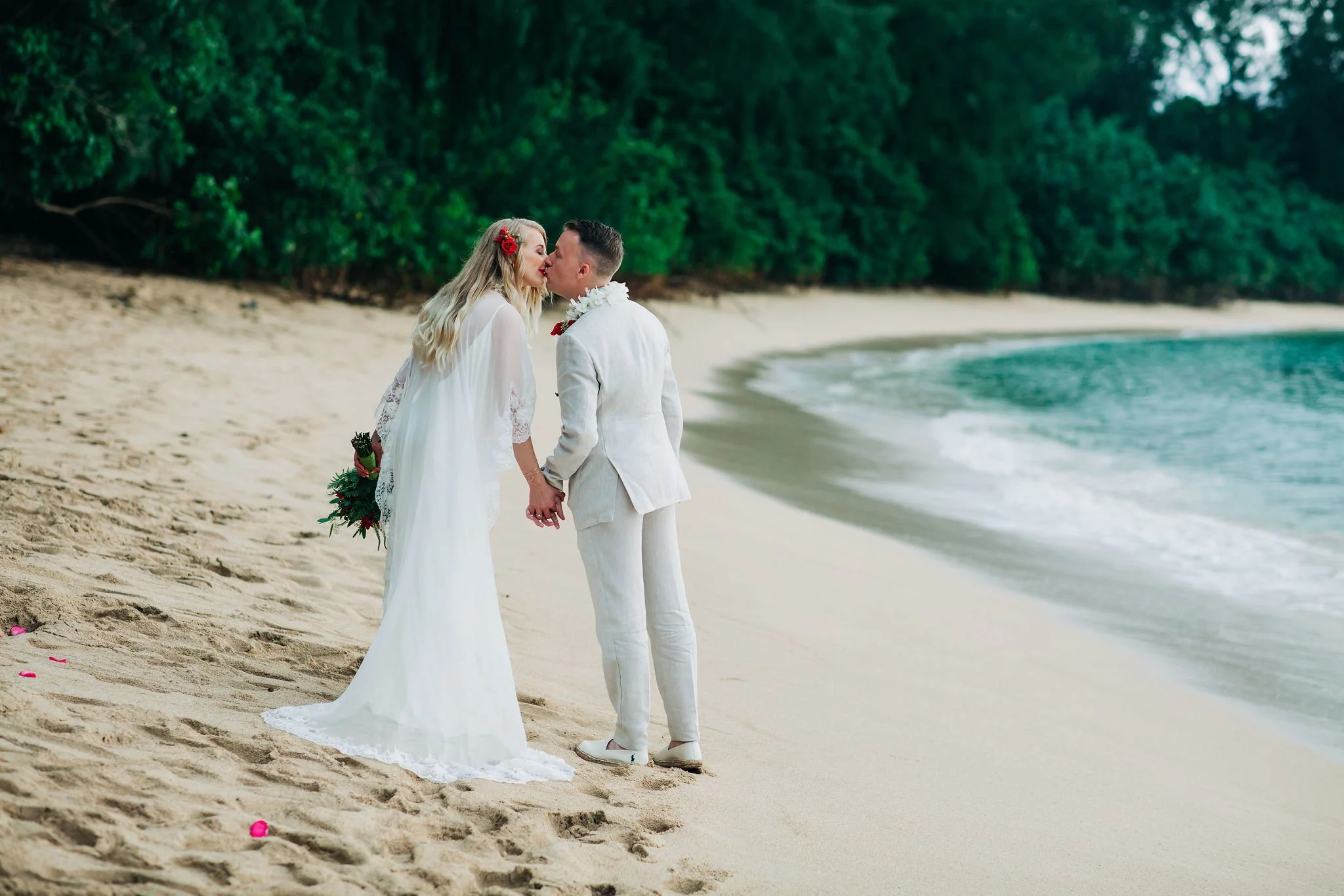 A bride and groom kissing on a beach with green trees in the background, holding hands, with the bride in a white wedding dress and the groom in a white suit.