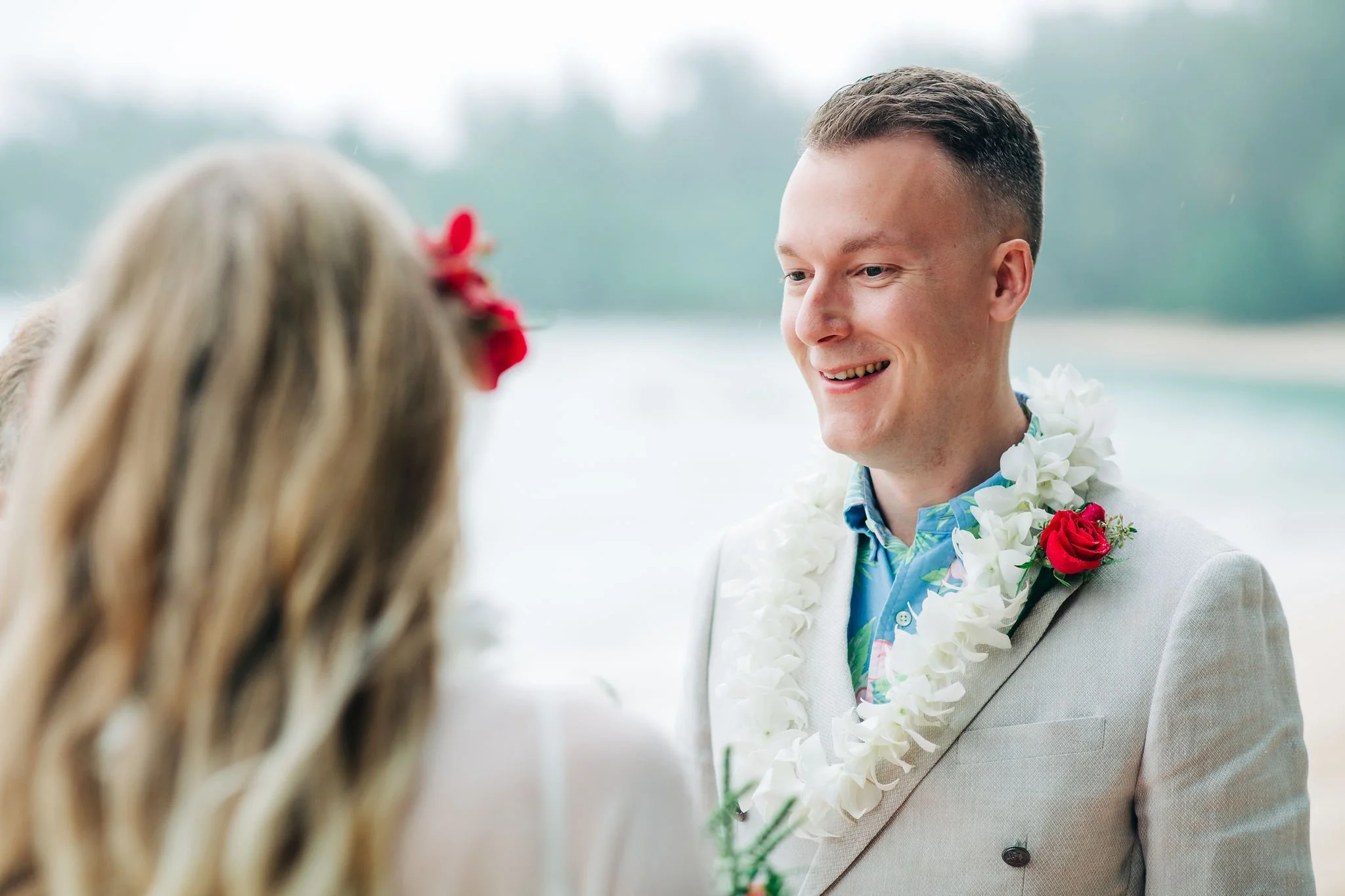 A groom standing outdoors at a beach wedding, smiling at the bride, wearing a lei, a floral shirt, and a coral boutonniere.