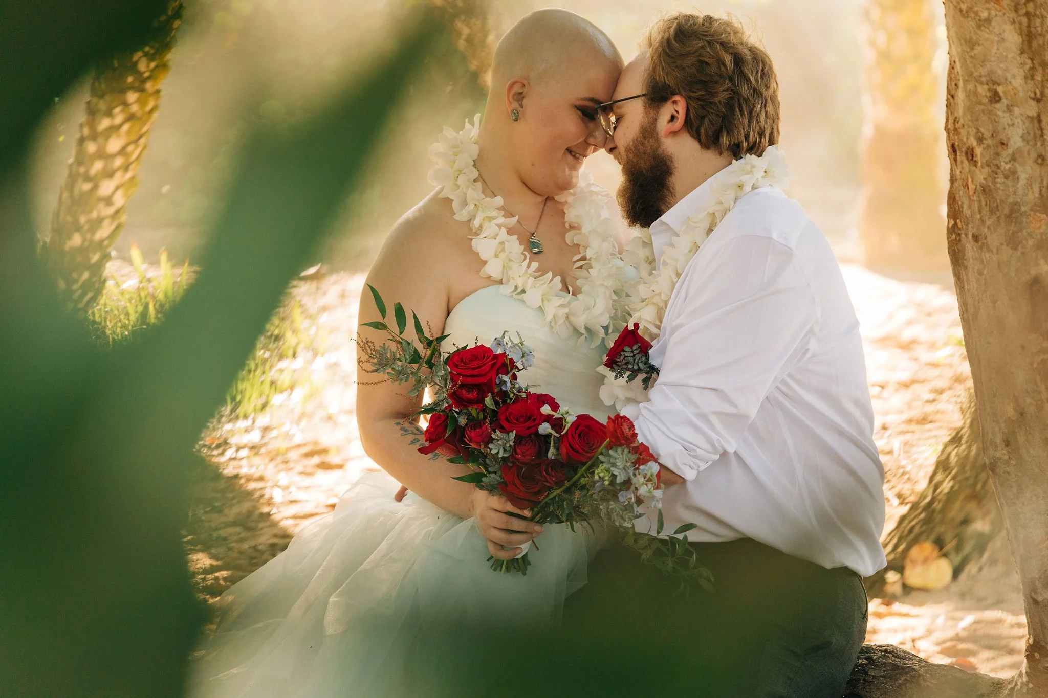 A couple dressed in wedding attire, smiling and touching foreheads, sitting close together outdoors, with the bride holding a bouquet of red roses and wearing a white dress and floral necklace, surrounded by trees and soft sunlight.