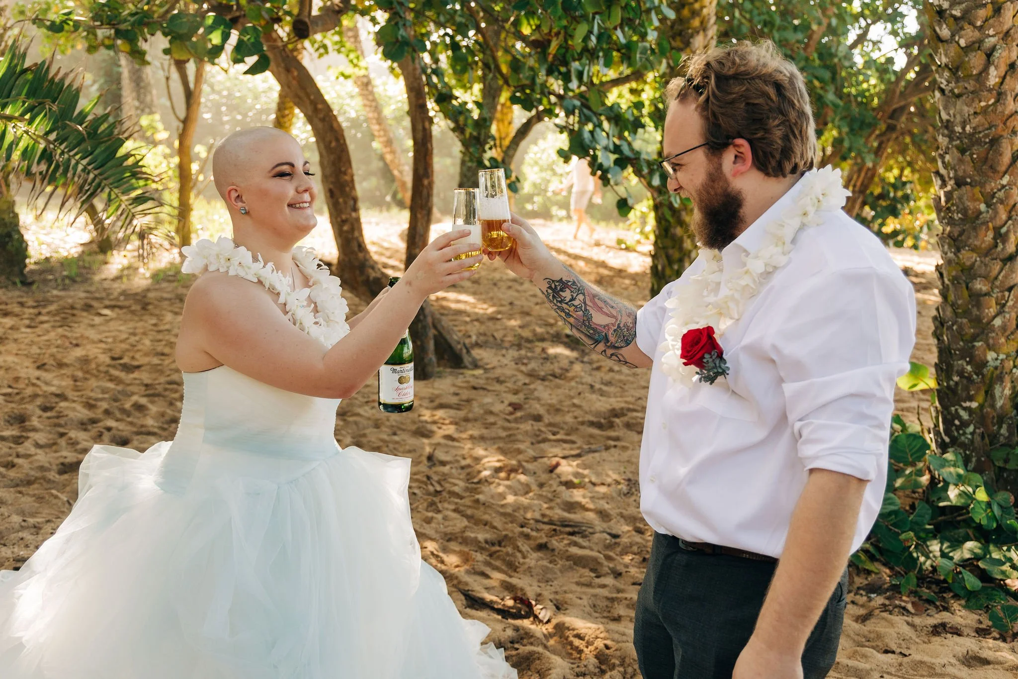 A bride with a shaved head wearing a white wedding dress and a groom with glasses and a beard wearing a white shirt and dark pants, toasting with drinks in an outdoor setting surrounded by trees.