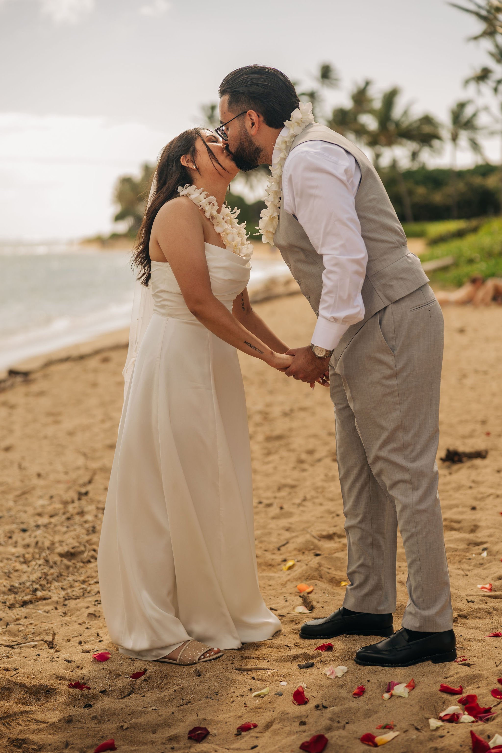 A couple dressed in wedding attire share a kiss while holding hands on a sandy beach with scattered flower petals, palm trees, and an ocean in the background.