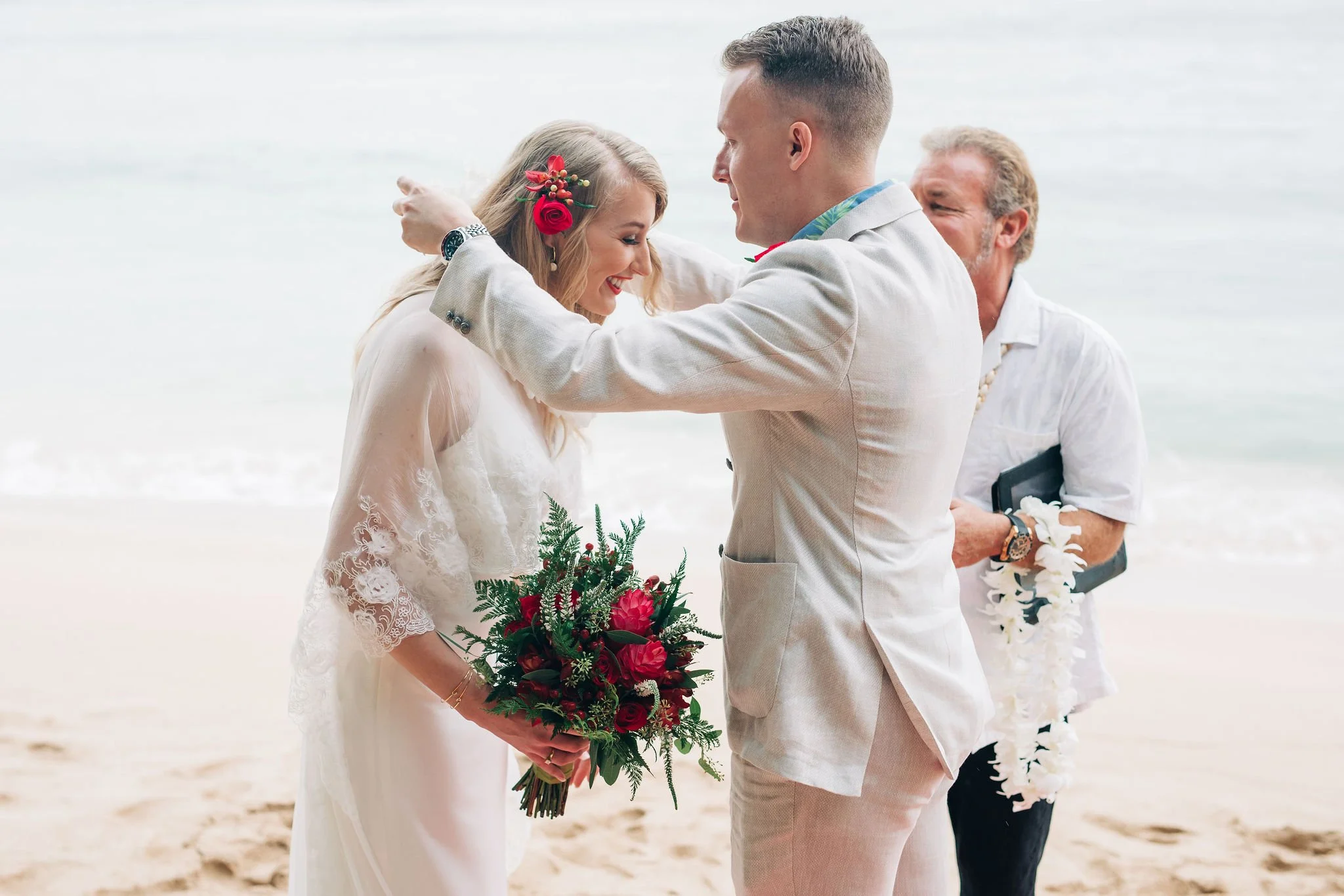 A couple in wedding attire sharing a moment on the beach, with an officiant preparing for the ceremony, during daytime.