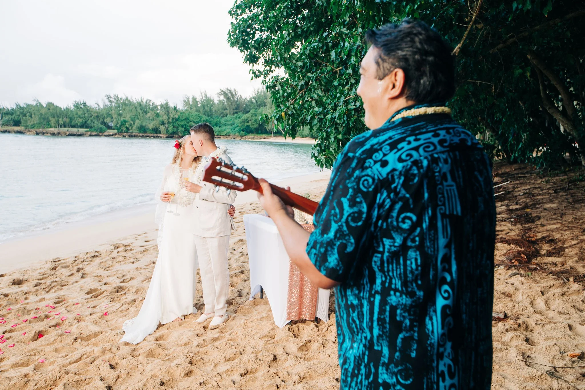 A couple in wedding attire sharing a kiss on the beach, with a musician playing guitar nearby, right next to a tree and water.