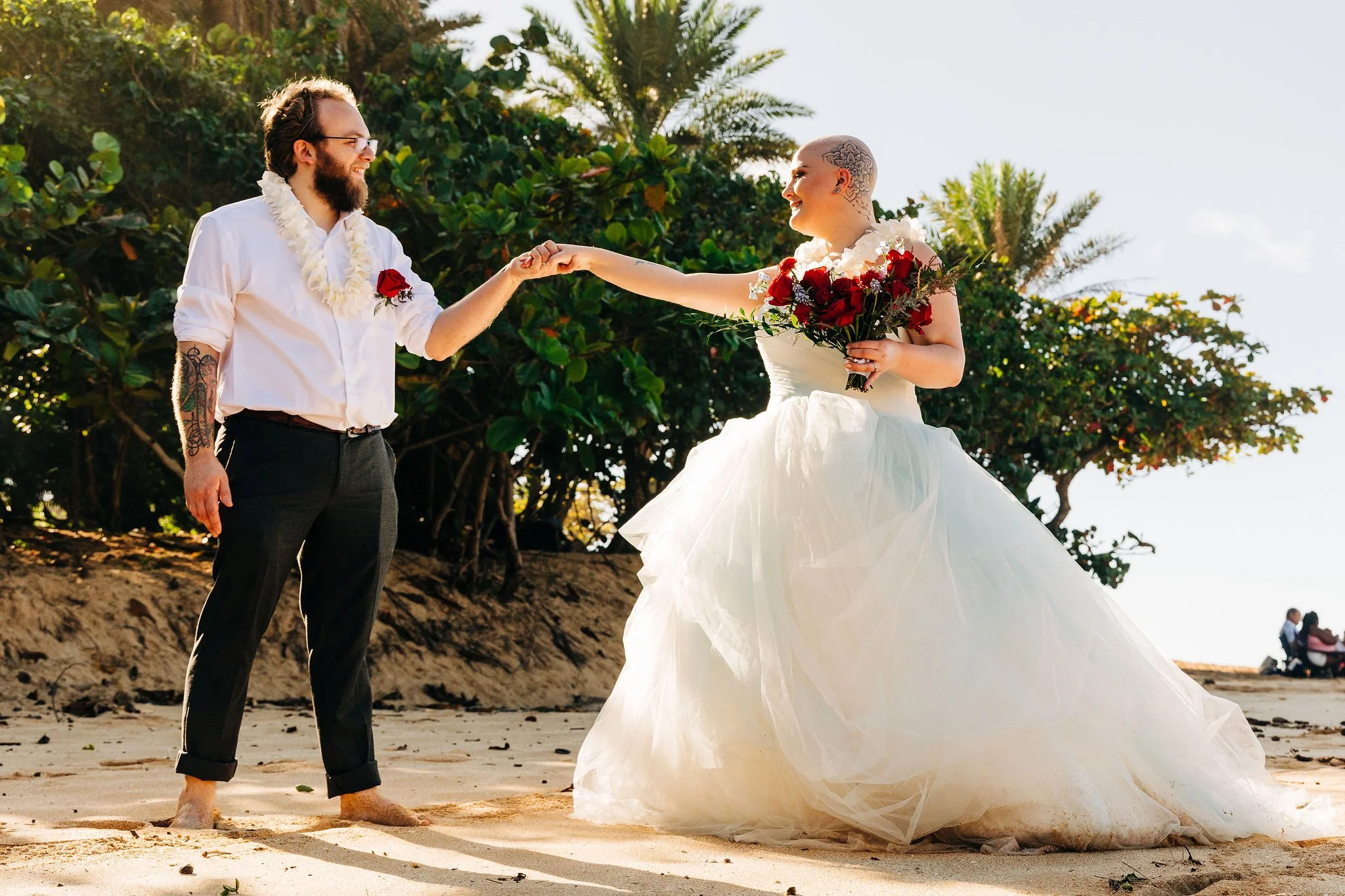 A couple getting married on a beach, the bride in a white wedding gown holding a bouquet of red and white flowers, and the groom in a white shirt and black pants, both smiling and holding hands.