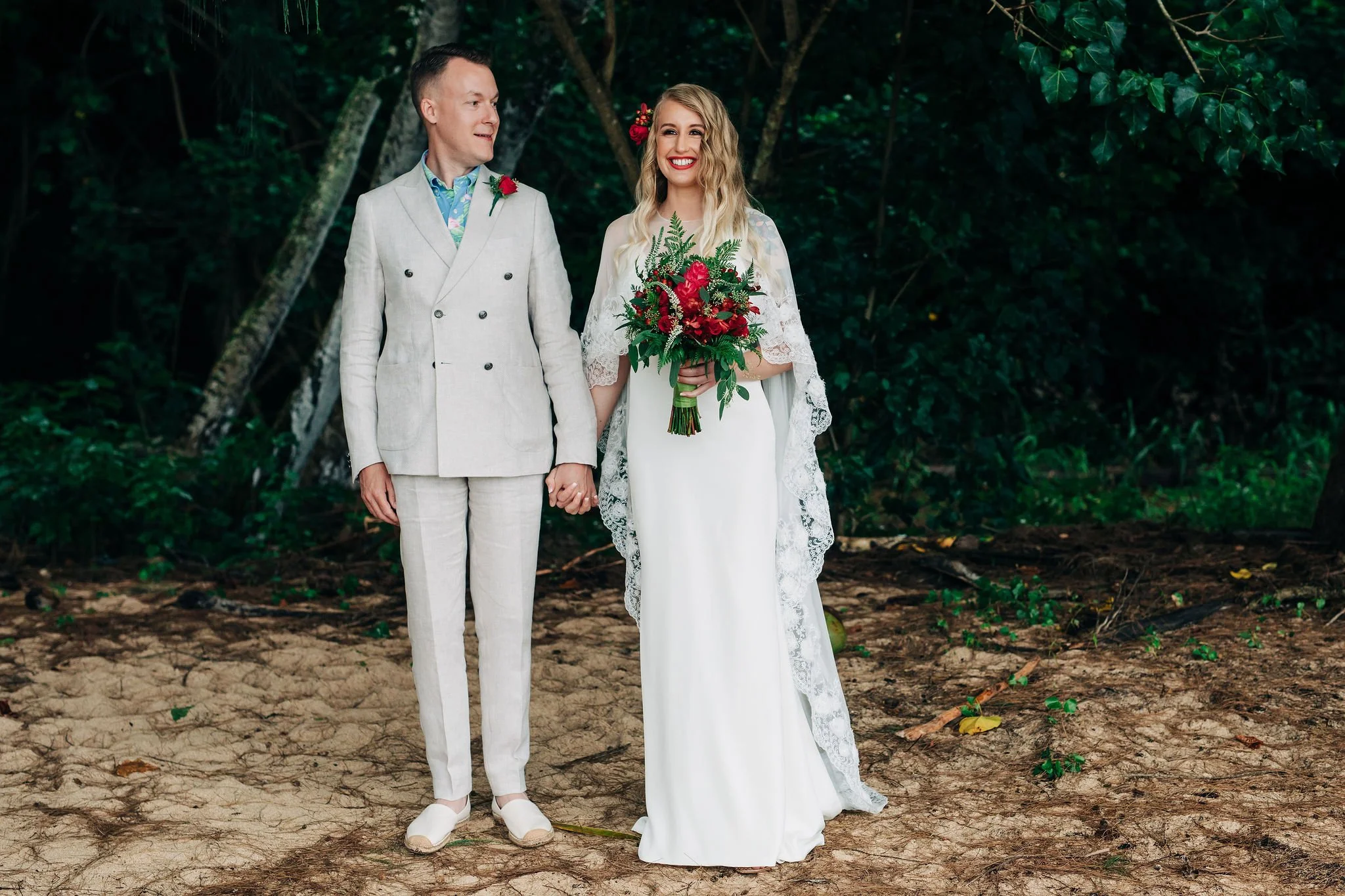 A bride and groom standing hand in hand in a wooded outdoor setting, dressed in wedding attire. The bride is holding a bouquet of red flowers and greenery, wearing a white gown with lace details and a long lace cape, and has a flower in her hair. The