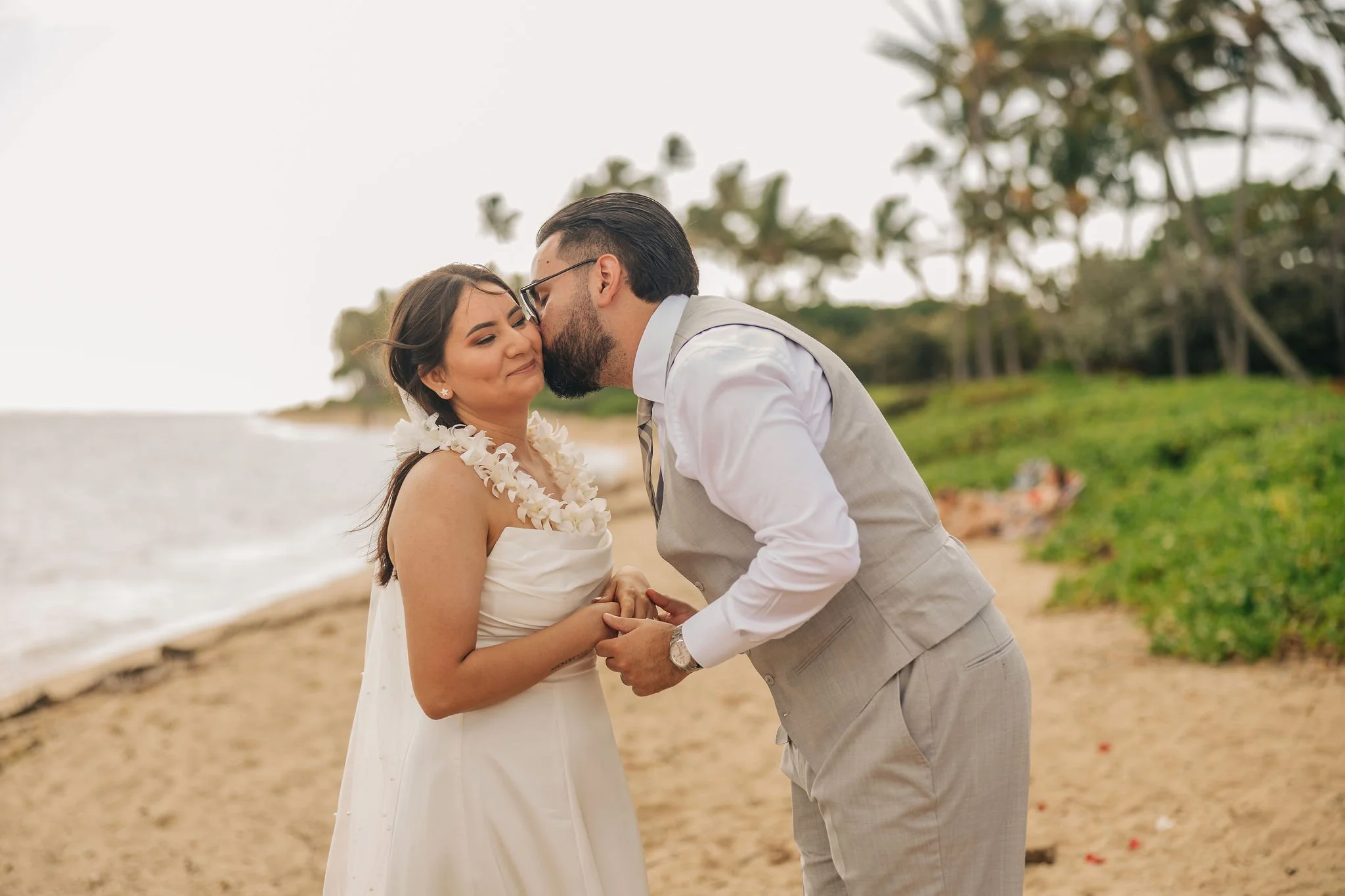 A couple dressed in wedding attire sharing an intimate moment on a beach, with the woman wearing a white dress and a floral necklace, and the man in a light gray suit, holding hands with the ocean and green trees in the background.