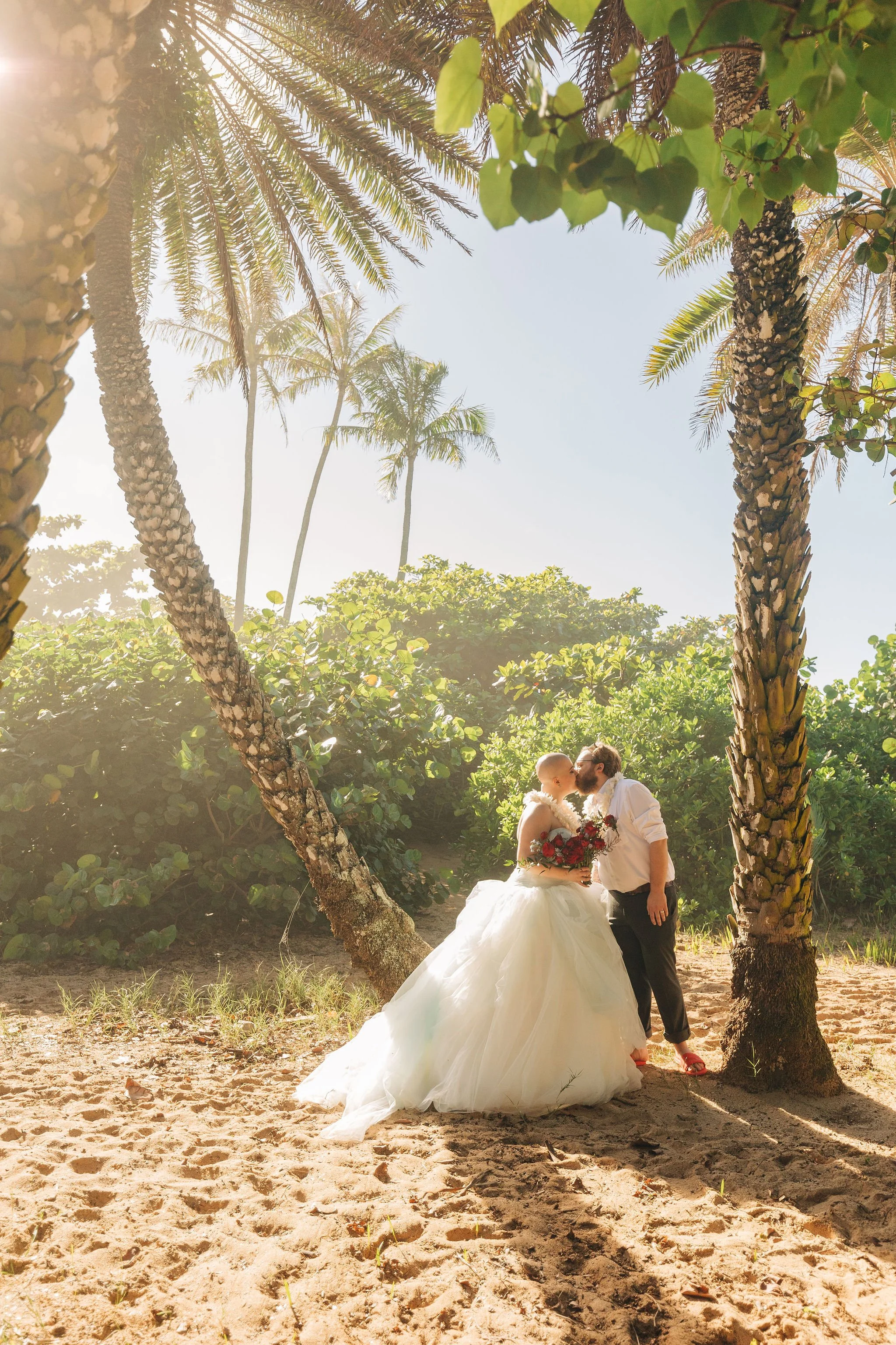 A wedding couple standing on sand beneath palm trees, the bride in a white wedding gown holding a bouquet, and the groom in black pants and a white shirt, kissing in a tropical outdoor setting.