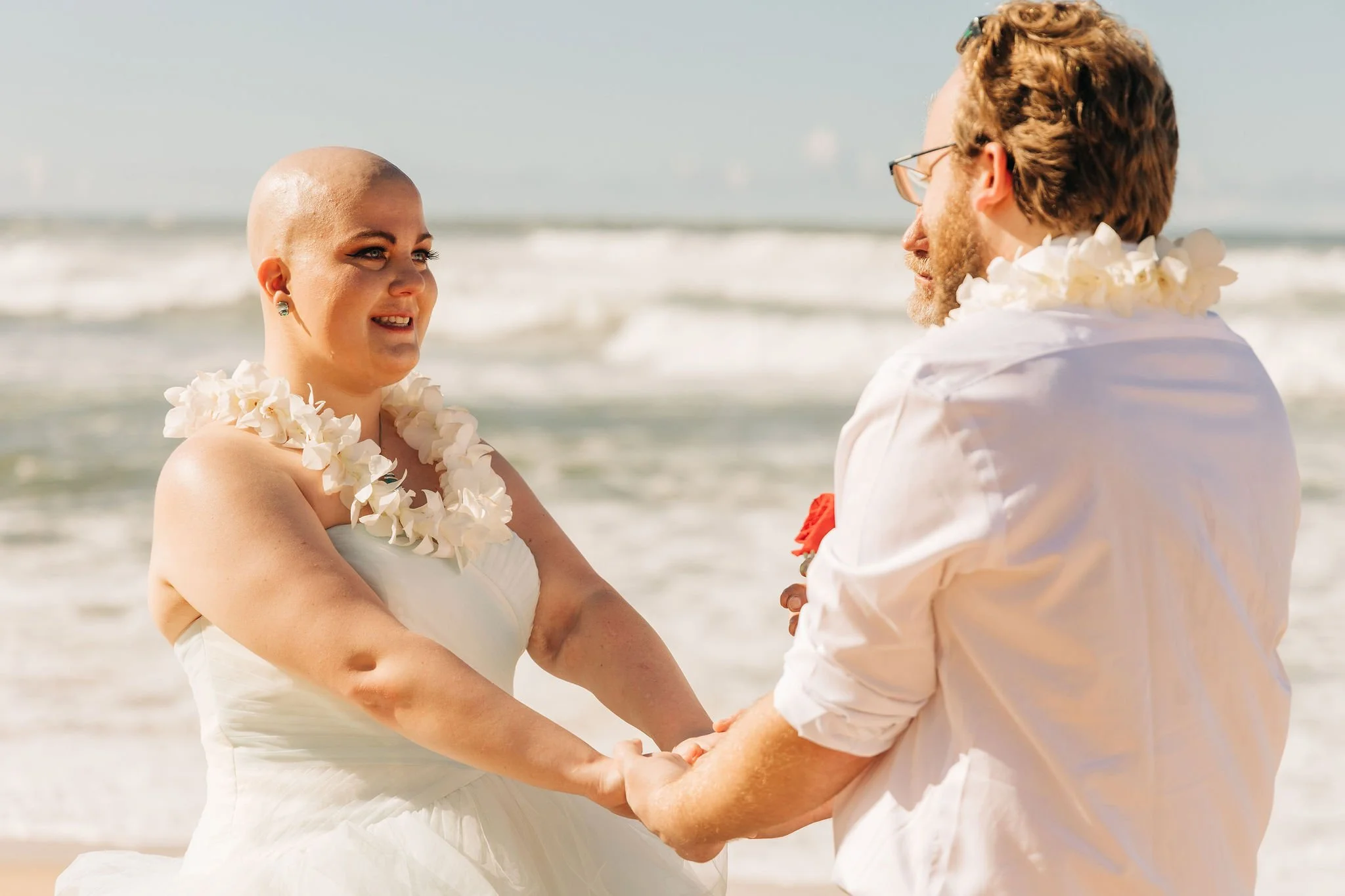 A couple gets married on the beach, holding hands and looking at each other, with the ocean in the background. The bride is wearing a white dress and a flower lei, and the groom is dressed in a white shirt, also with a flower lei.