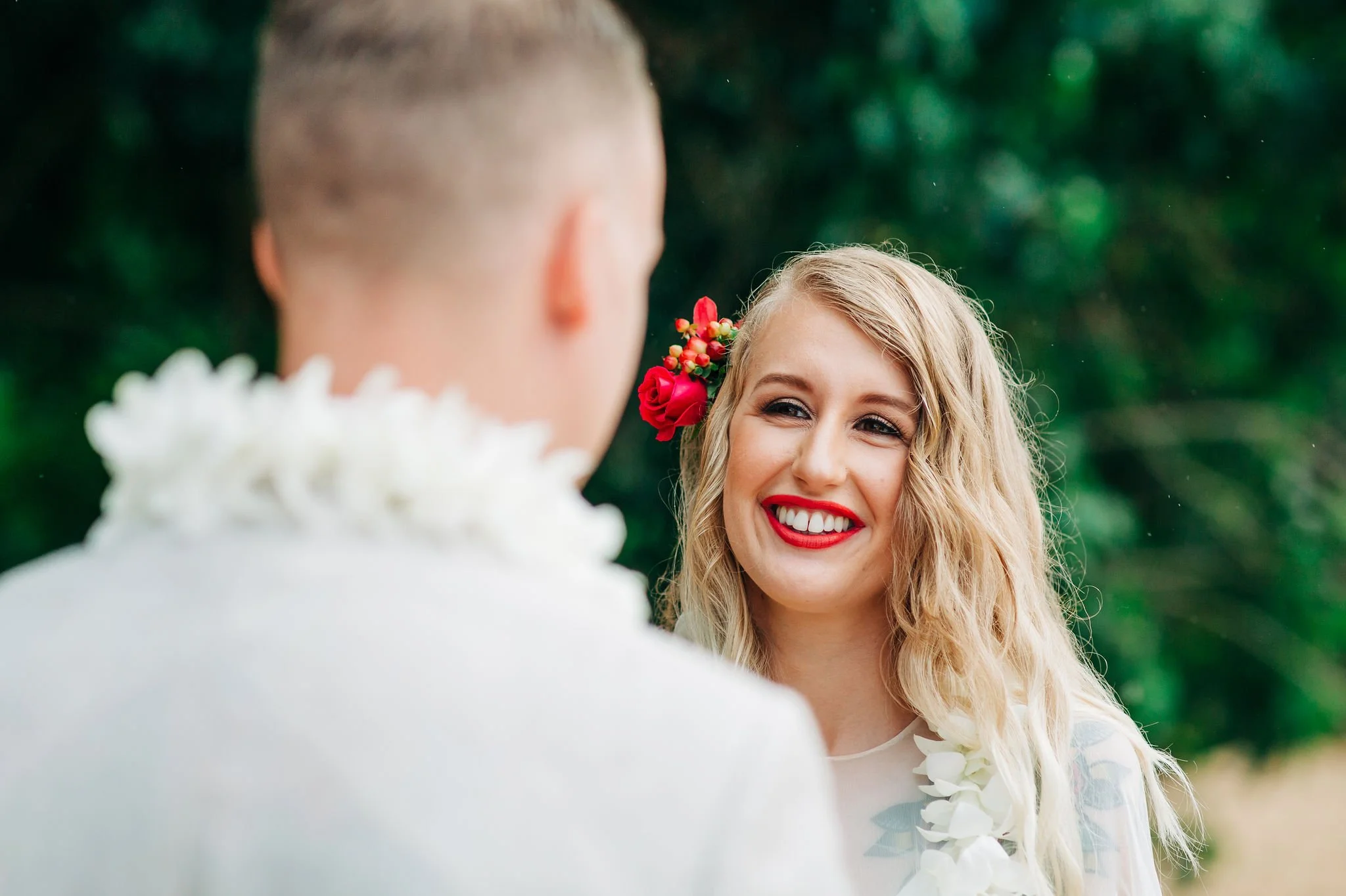 A woman with wavy blonde hair and bright red lipstick smiling at a man whose back is to the camera. She has a red and pink flower in her hair and is wearing a white outfit with white floral accessories. The background is green and blurred, indicating