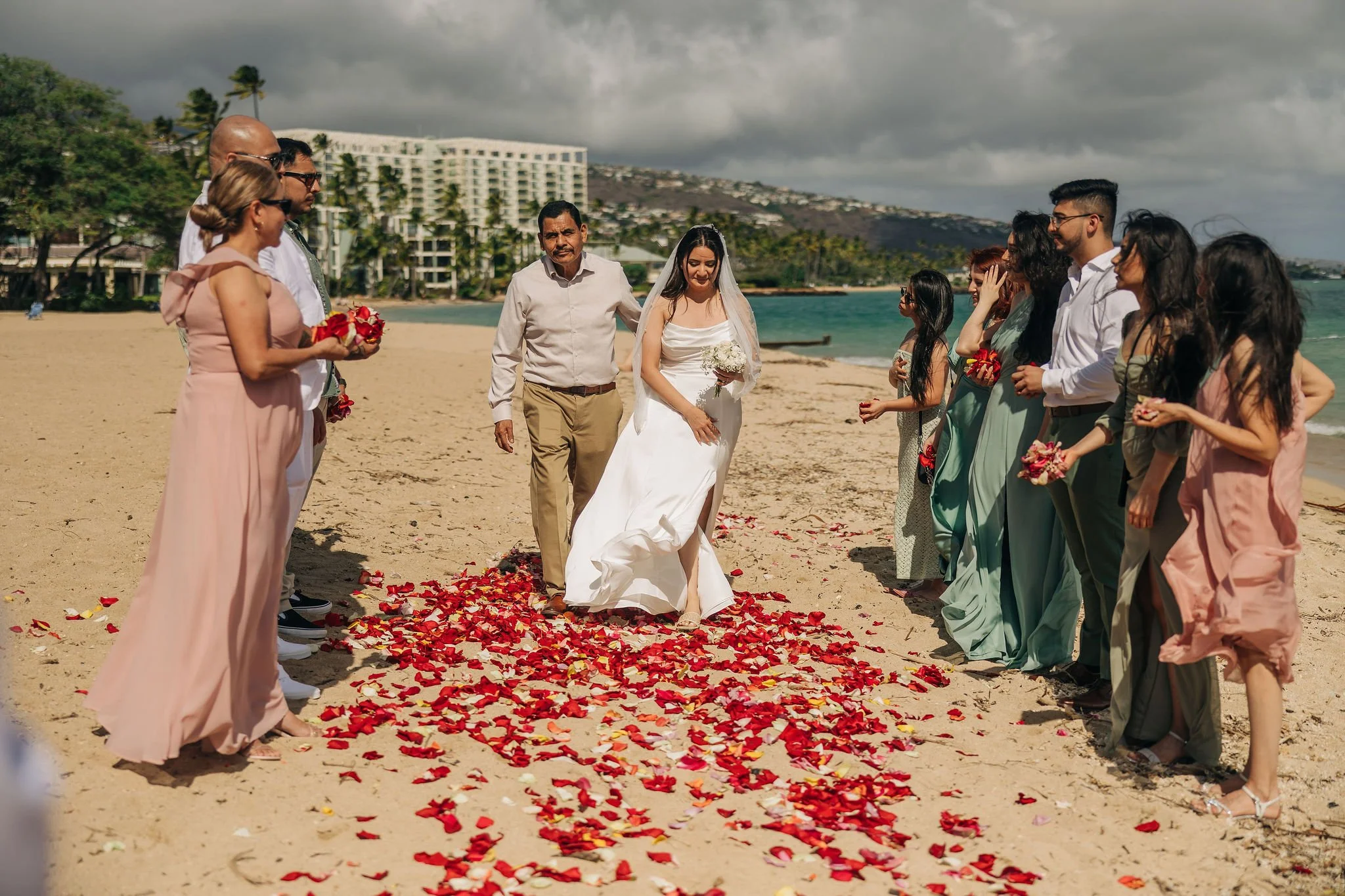A bride and groom walking on a beach with flower petals on the sand, surrounded by family and friends during a wedding ceremony under a cloudy sky.