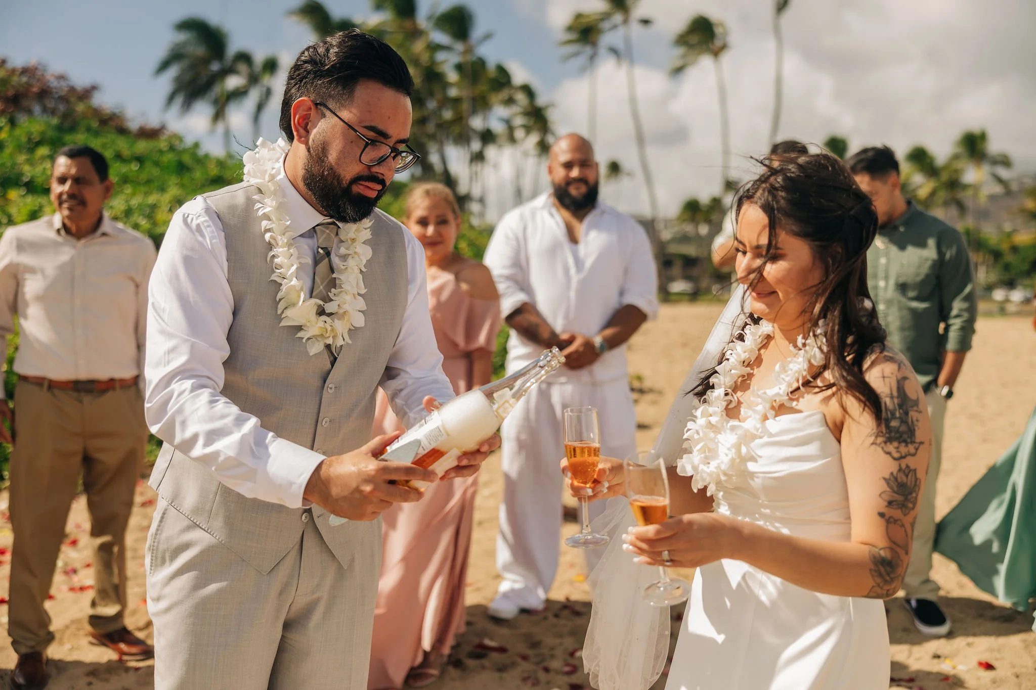 A wedding ceremony on a beach with a man in a beige vest and glasses pouring champagne for a woman with tattoos in a white dress, surrounded by friends and palm trees.