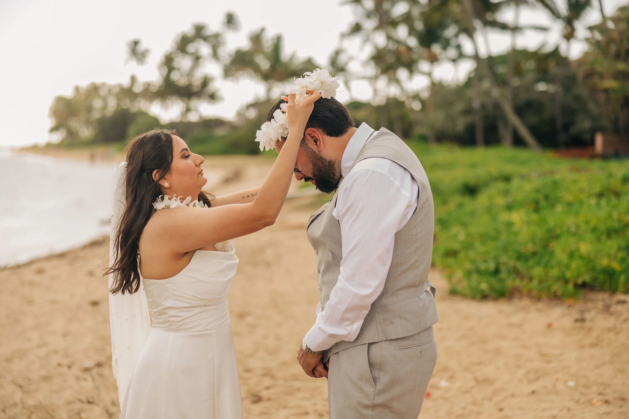 A couple on the beach during a wedding, with a woman placing a flower crown on a man’s head. The woman is wearing a white dress with a floral necklace, and the man is dressed in a light gray vest and pants.