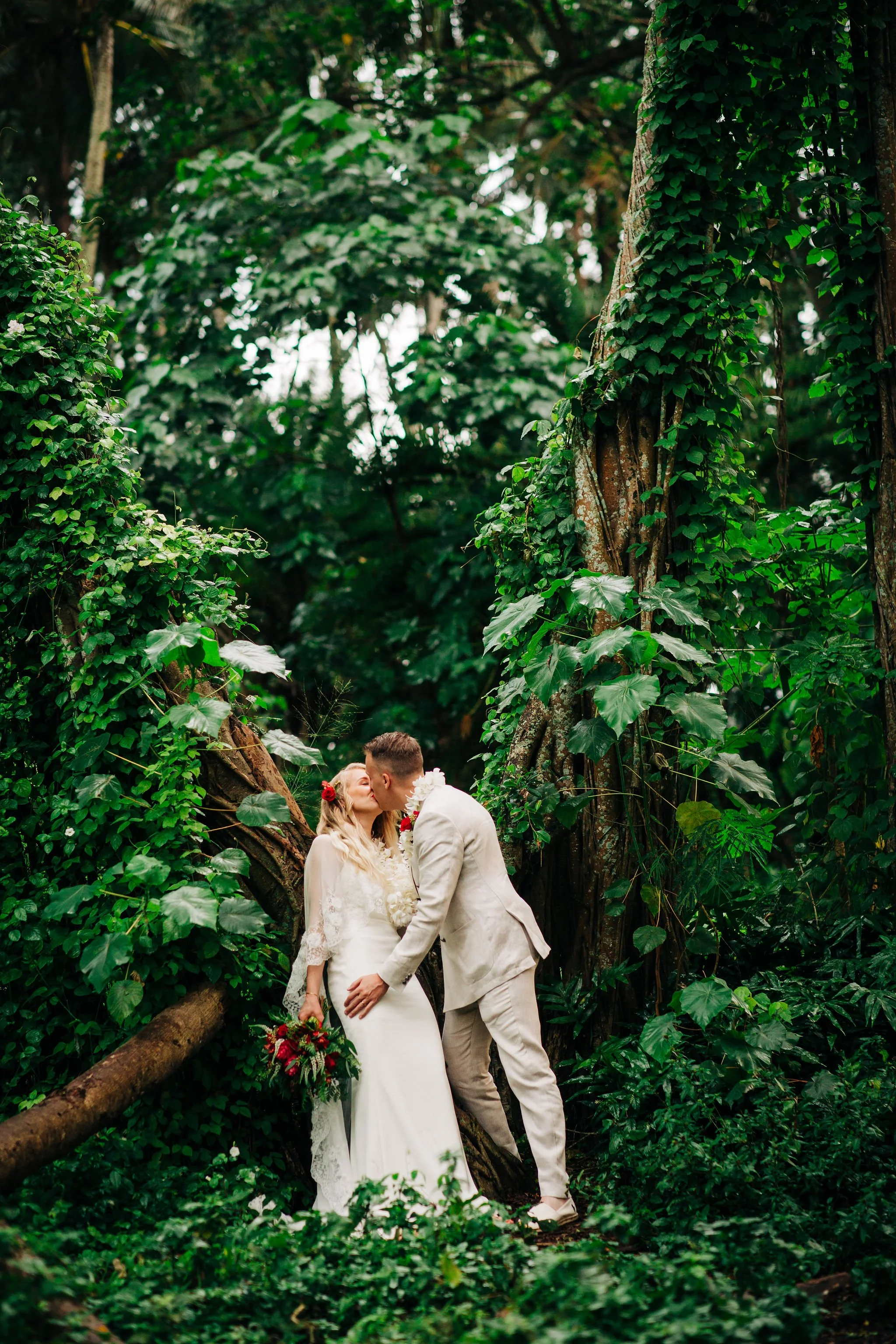 A bride and groom share a kiss in a lush, green forest surrounded by dense foliage and tall trees.