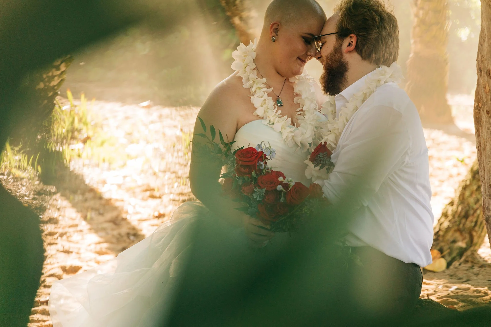Couple in wedding attire sitting closely together outdoors, surrounded by trees and sunlight, with the bride holding a bouquet of red roses.