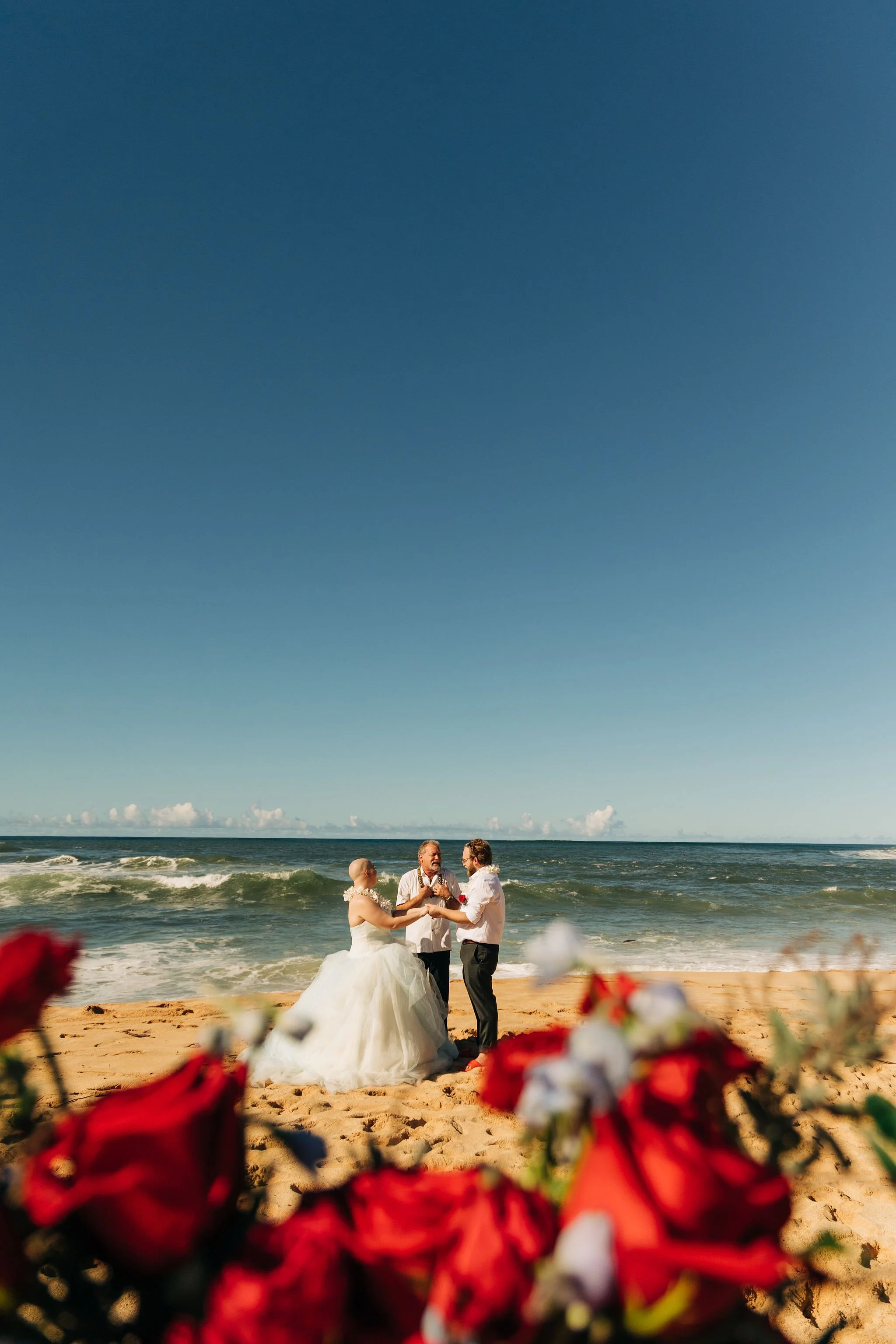 A wedding ceremony taking place on a beach with the ocean and blue sky in the background, featuring a bride in a white gown, a groom in a white shirt and dark pants, and an officiant.