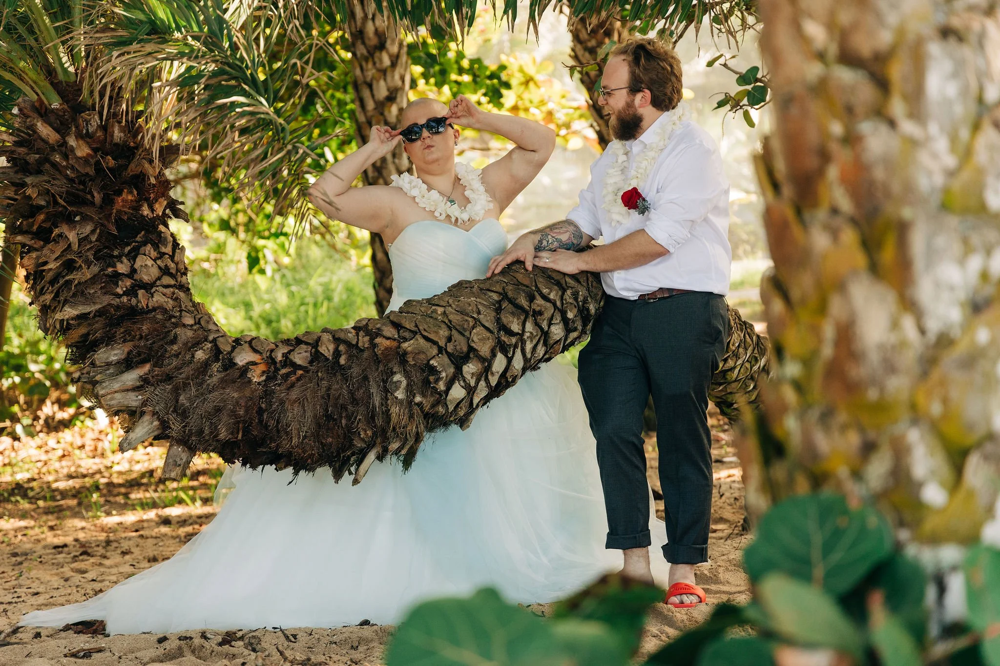A couple dressed in wedding attire poses on a large palm tree in a tropical outdoor setting. The woman is wearing a strapless white wedding dress, sunglasses, and a white floral necklace, while the man is dressed in a white shirt with rolled-up sleev